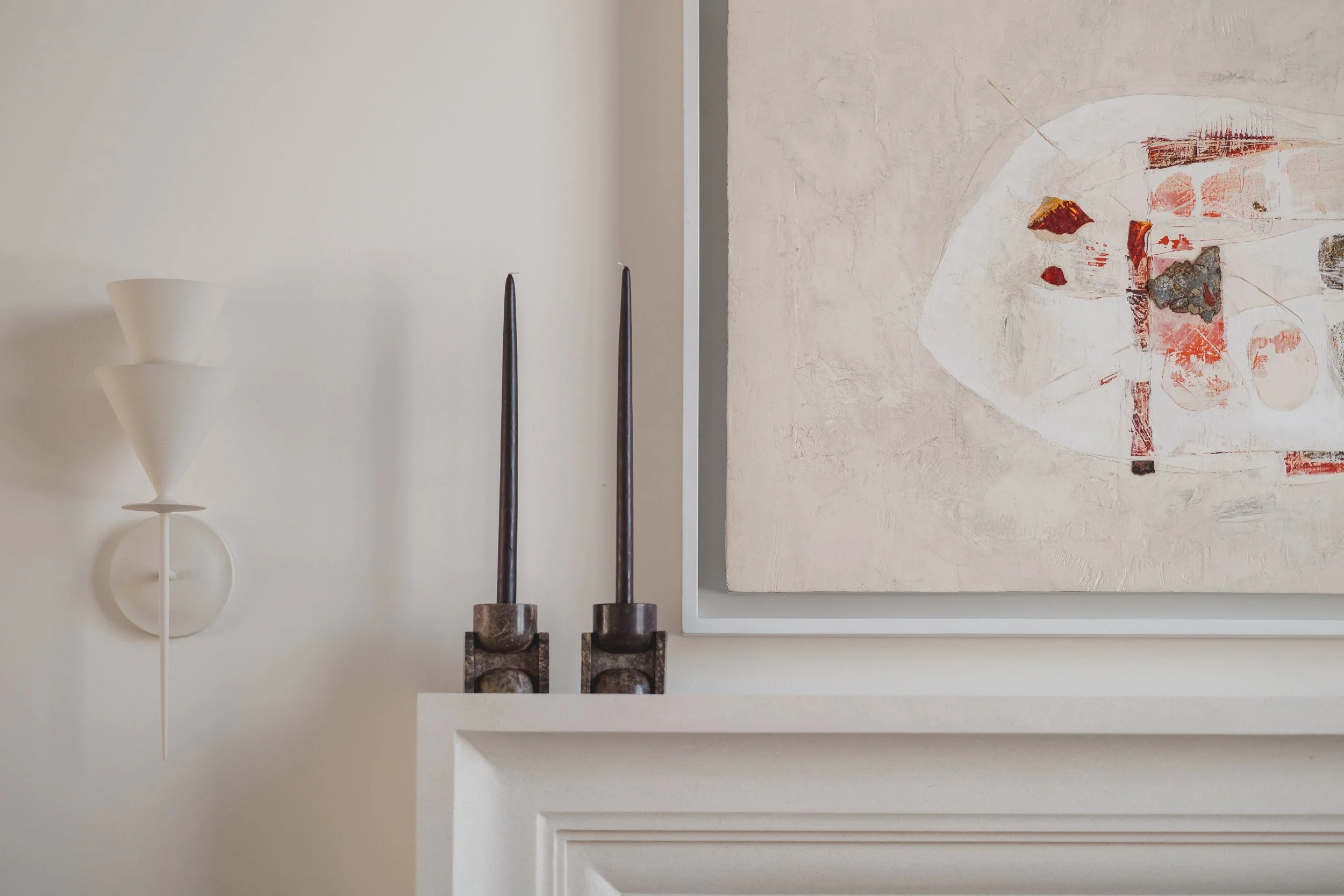 Interior decor with two black candlesticks, abstract artwork, a white wall-mounted lamp, and part of a white mantelpiece.