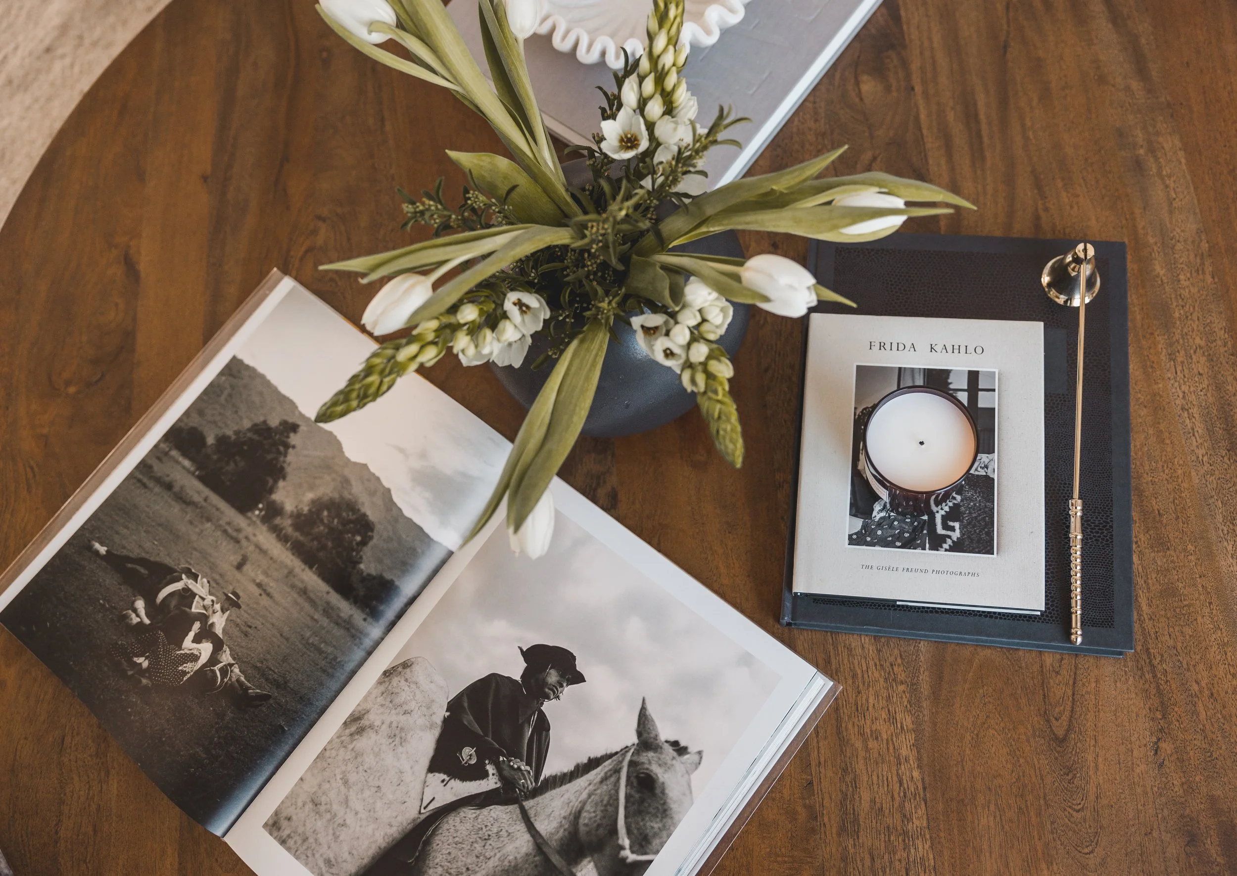Open photo album with black and white photographs of people, a vase with white flowers on a wooden table, a book about Frida Kahlo with a candle on top, and a metallic pen resting on a black notebook.