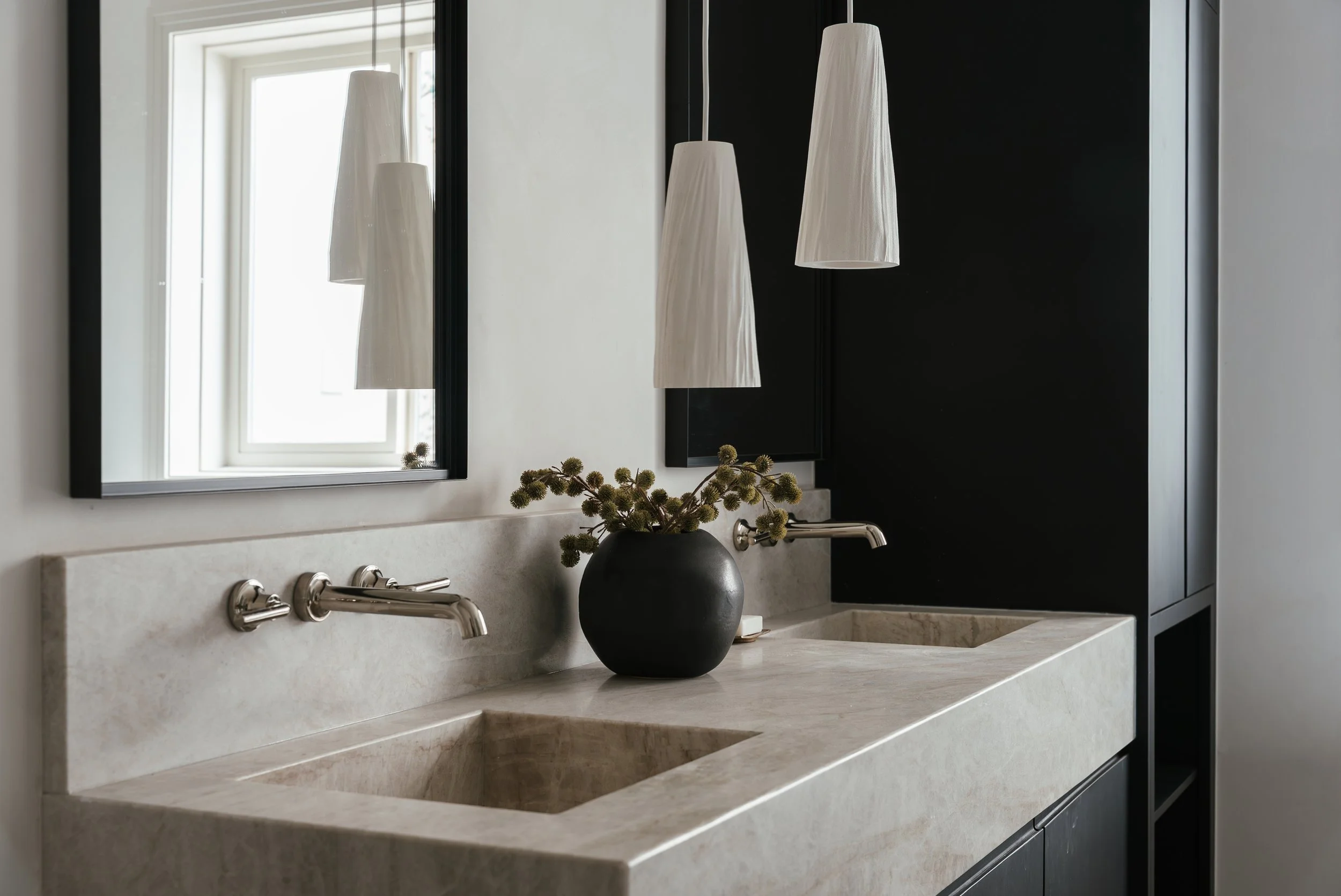 A modern bathroom with a double beige marble sink, black cabinets, large mirrors, white pendant lights, a window, and a round black vase with green plants.