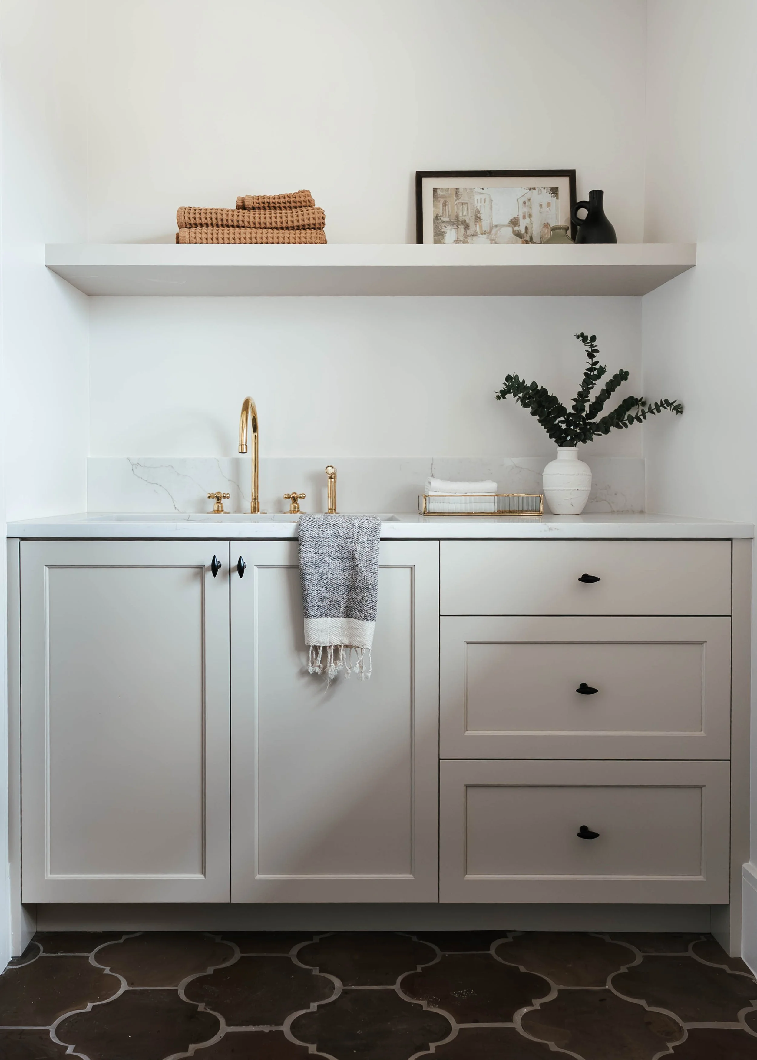 A minimalist bathroom vanity with white cabinets, a white marble countertop, black handles, and a gold faucet. There is a gray and white towel hanging on the sink, a Moroccan-style tile floor, a white vase with green foliage on the right, gold tray w