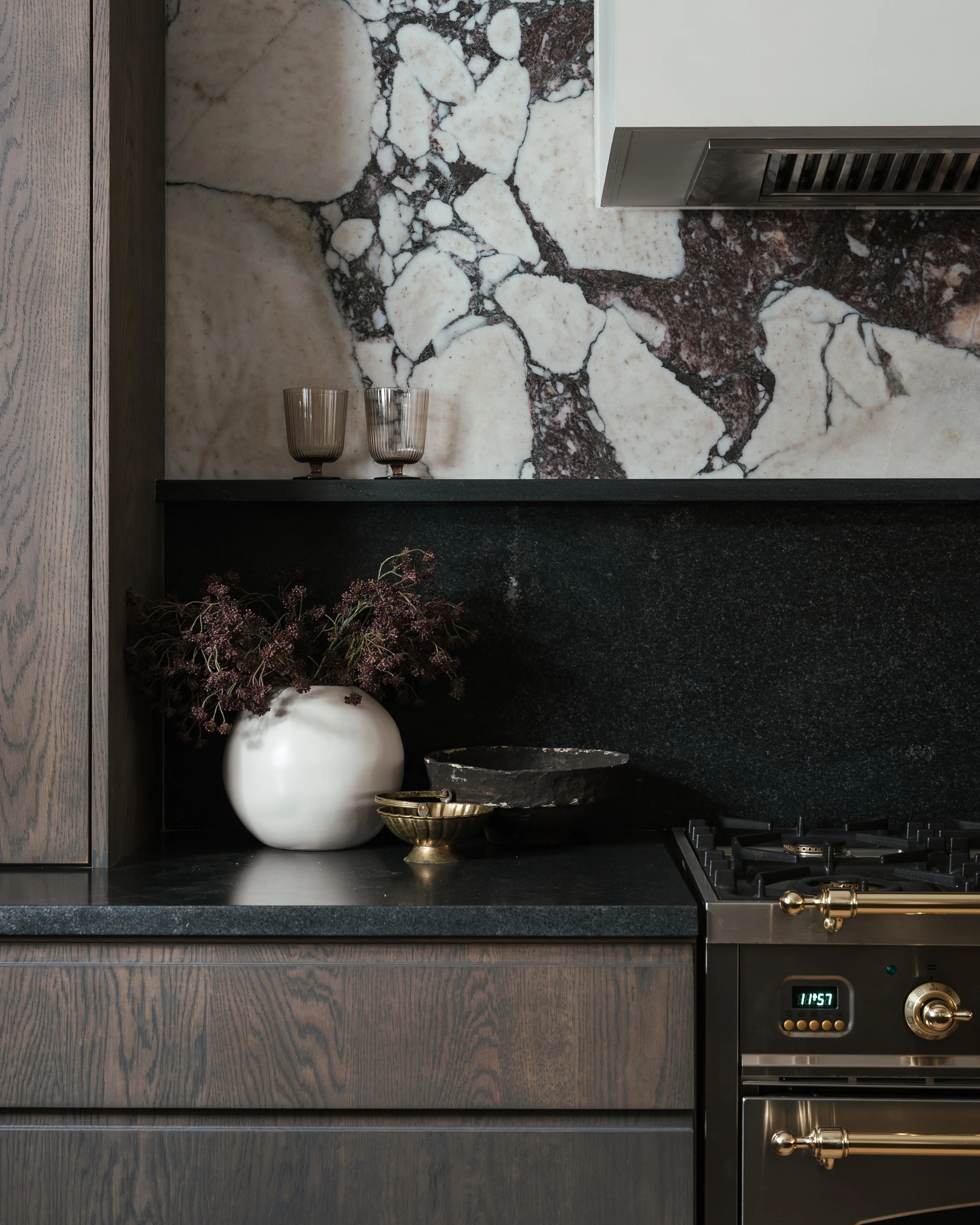 Close-up of a modern kitchen with dark wood cabinets, black countertop, marble backsplash, and decorative items including a white vase with purple flowers, glasses, and bowls, along with a stove and oven.