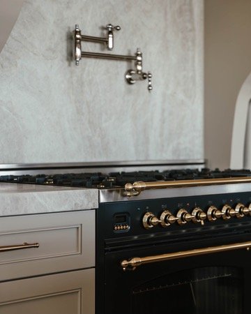 Close-up of a modern kitchen stove with brass knobs and a gray countertop, with a metallic pot filler faucet mounted on a textured gray wall above.
