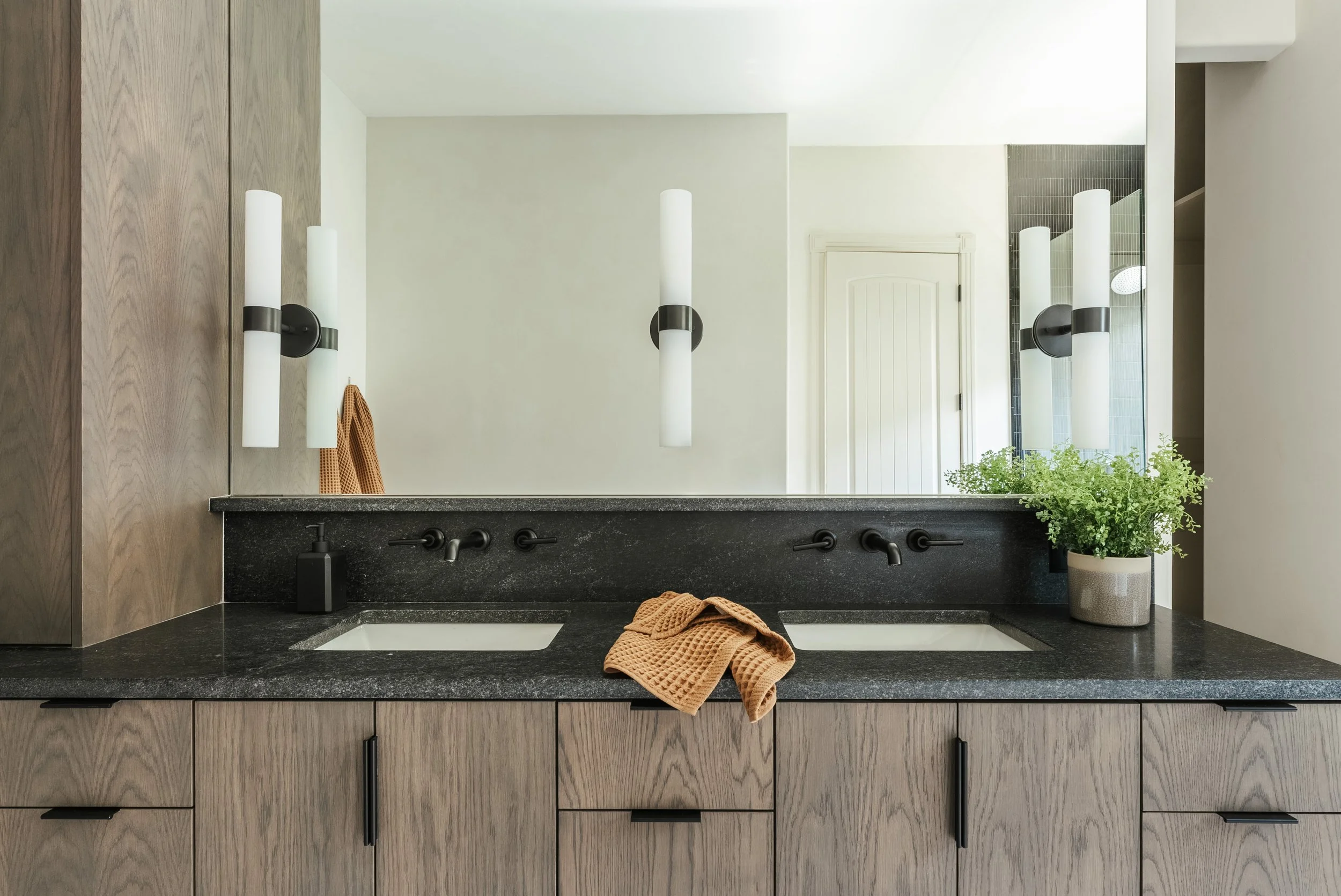 Modern bathroom vanity with dual sinks, black countertop, and wooden cabinets. A large mirror is above the sinks, with wall-mounted light fixtures on either side. A potted green plant is on the right, and a beige towel is draped over the counter.