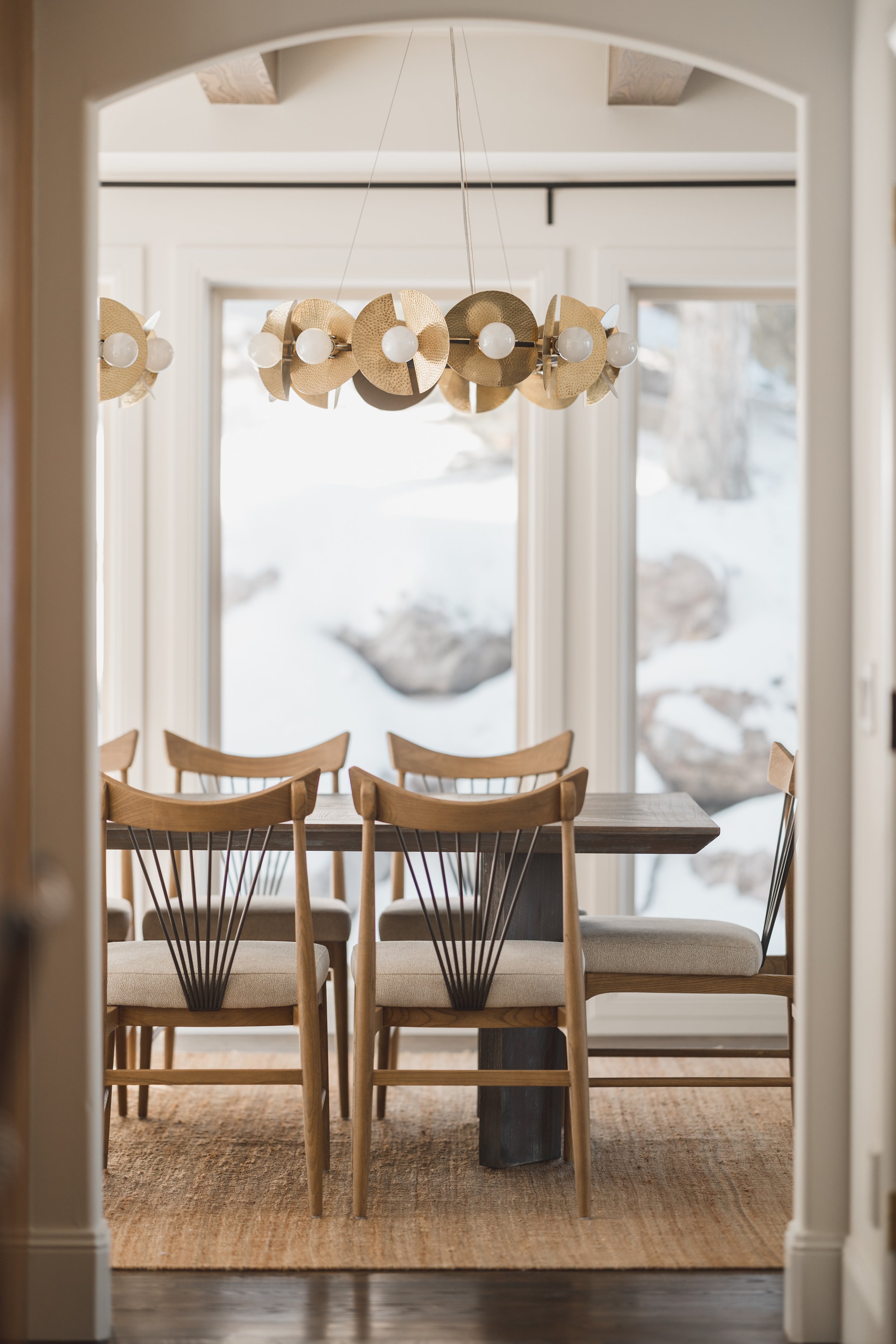 View through a doorway of a dining room with a wooden table, six wooden chairs with light-colored cushions, a modern chandelier with round light bulbs, and large windows showing a snowy outdoor landscape.