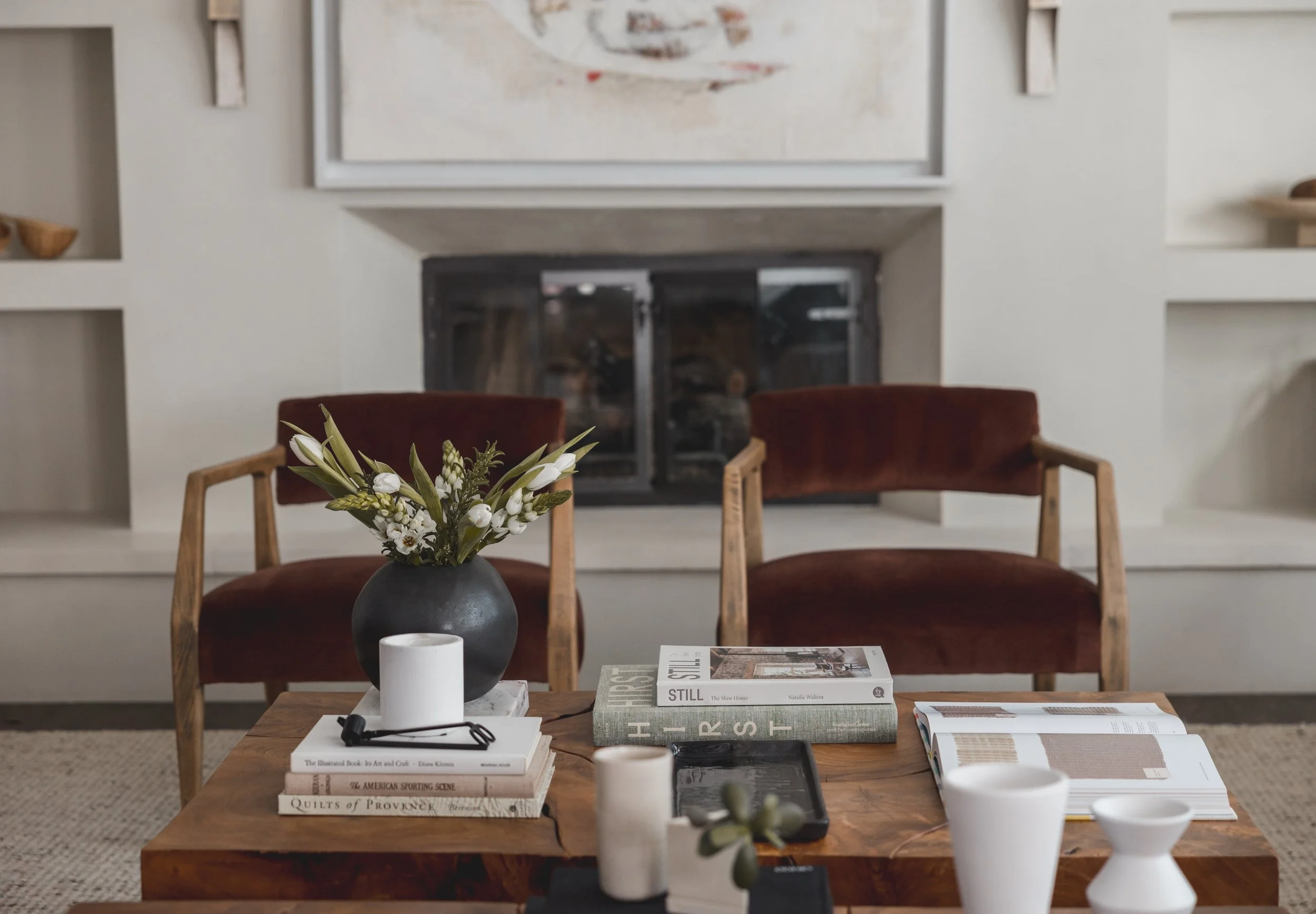 A cozy living room with a wooden coffee table, two burgundy armchairs, a fireplace, and a wall-mounted abstract painting. The table has a black vase with white flowers, candles, books, and a tray.