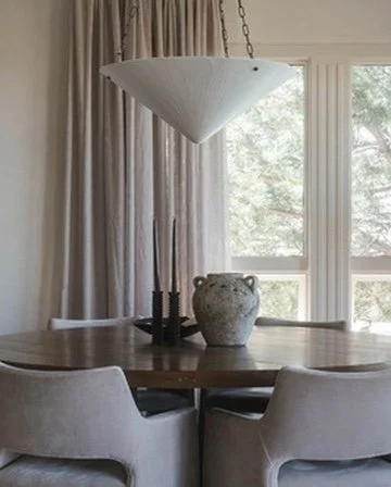 Dining room with a round wooden table, four chairs, a white pendant lamp, a decorative vase, and large windows with curtains showing trees outside.