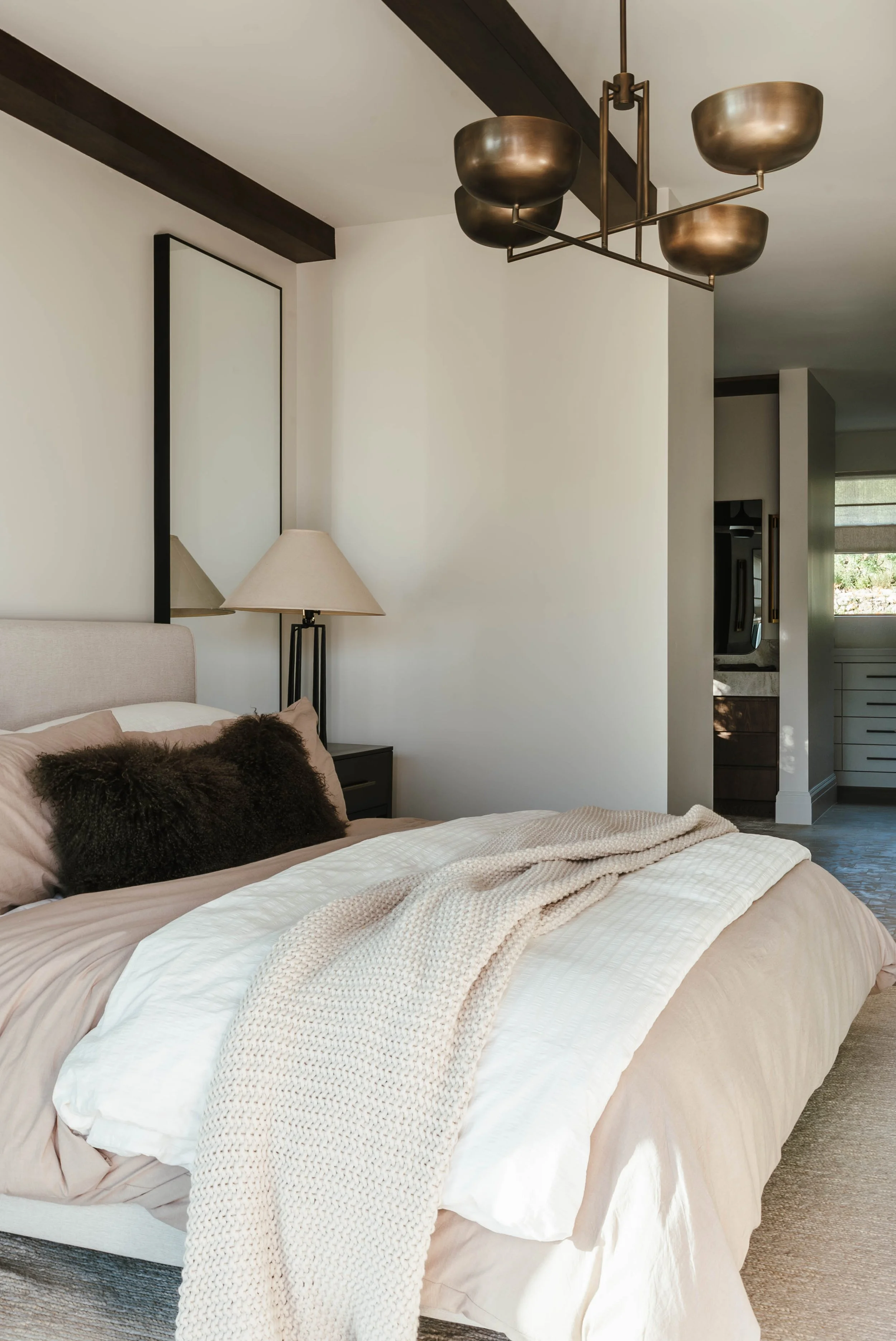 A neatly made bed with beige linens, a dark brown fluffy pillow, and a knitted beige blanket. A nightstand with a lamp is next to the bed, and there's a large rectangular mirror on the wall. A modern chandelier hangs from the ceiling, and part of the