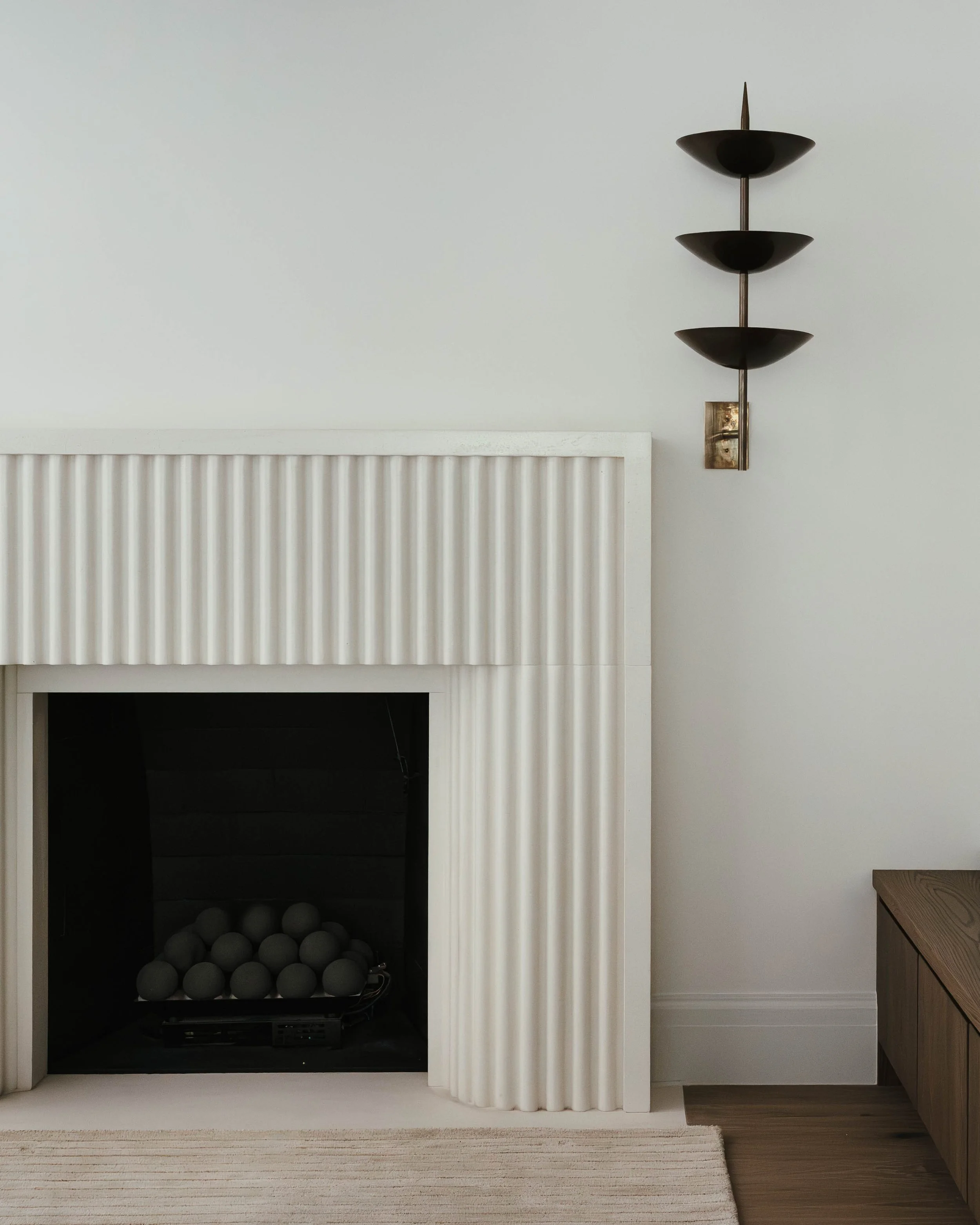 White fireplace with black spheres inside, modern wall sconce with three black semi-circular shades on a white wall, wooden cabinet on the right, striped rug on the floor.