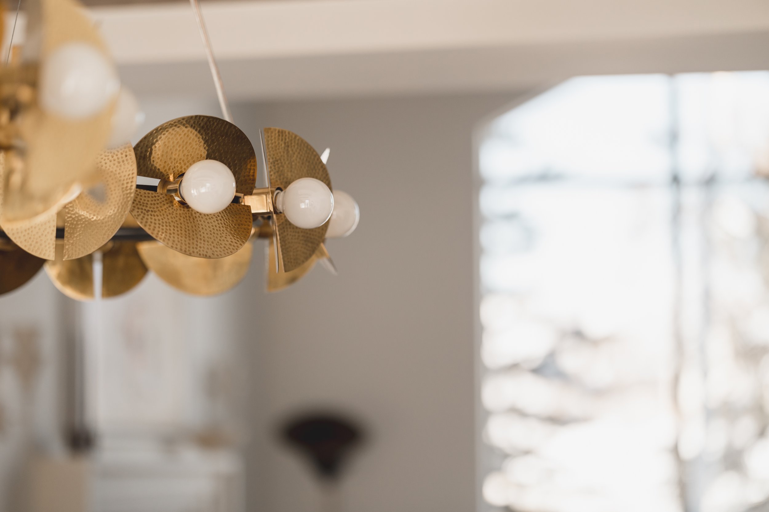A modern chandelier with gold-colored, perforated metal discs and white spherical light bulbs hanging from a ceiling, with a window in the background letting in natural light.
