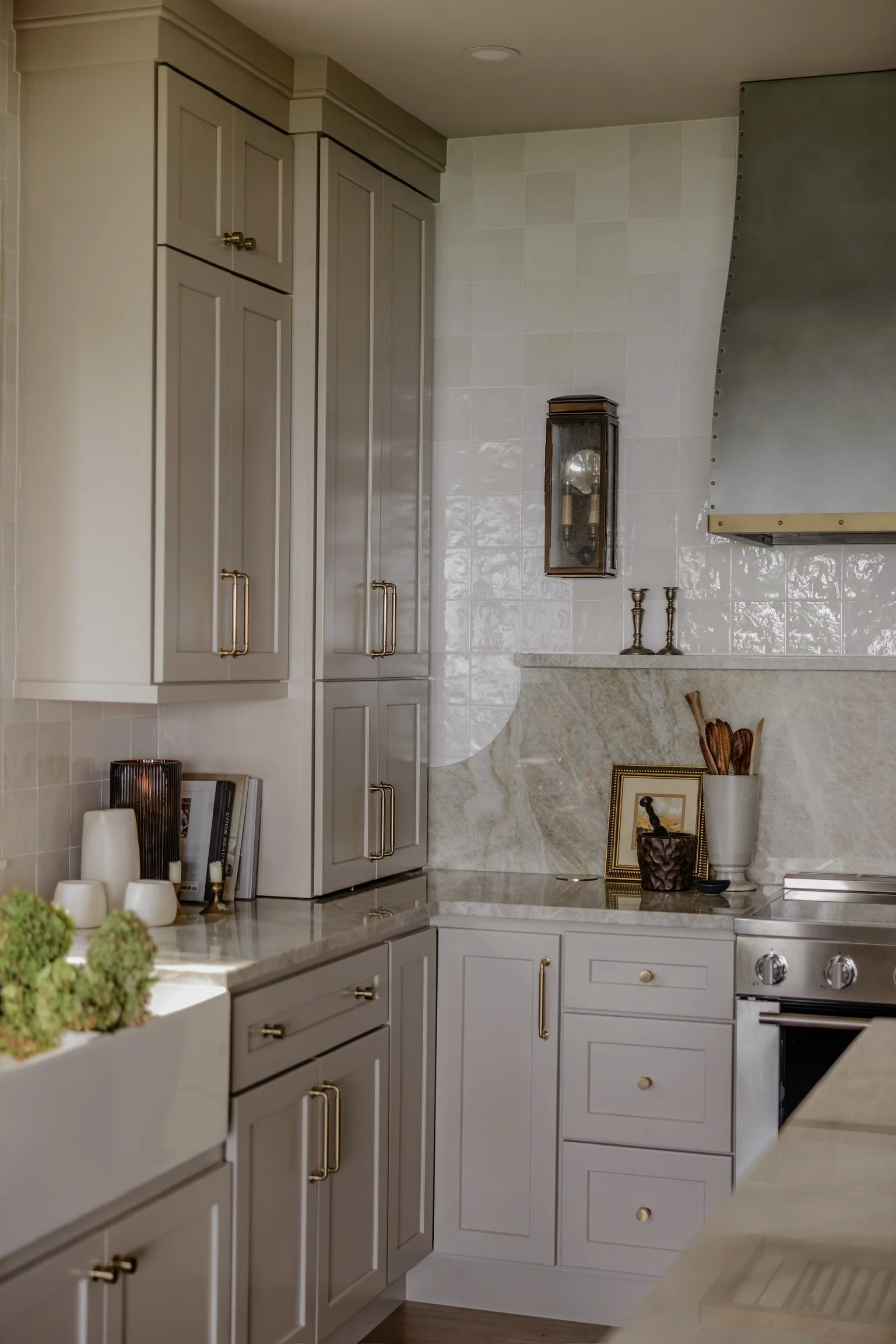 Kitchen with beige cabinets, marble countertops, and a stainless steel stove. Decor includes a framed picture, a white vase with wooden utensils, candlesticks, and books on the countertop. There is a textured marble backsplash and a wall-mounted ligh