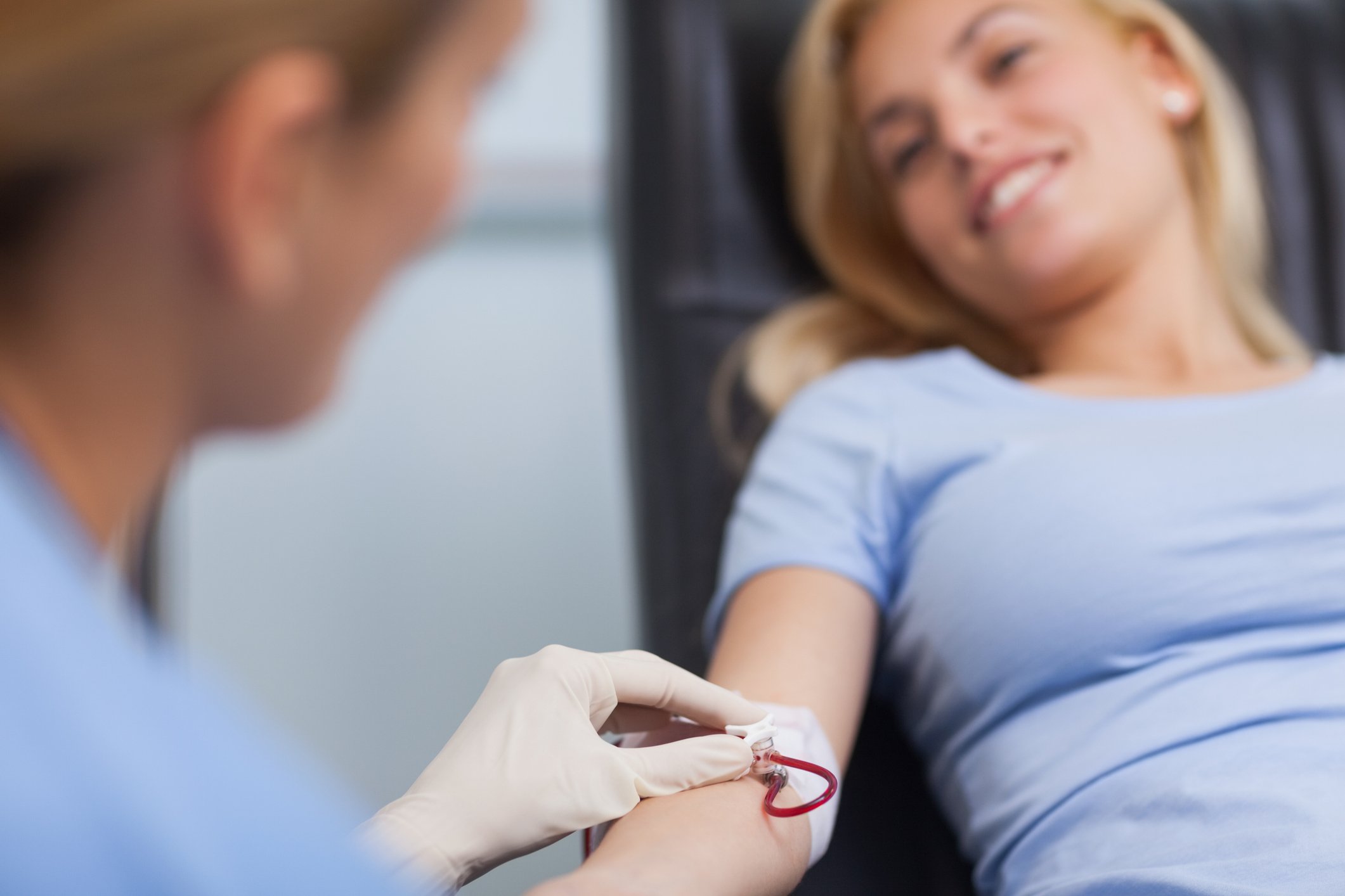A woman is sitting comfortably while a healthcare professional draws blood from her arm for a blood donation or blood test. The woman is smiling and relaxed.