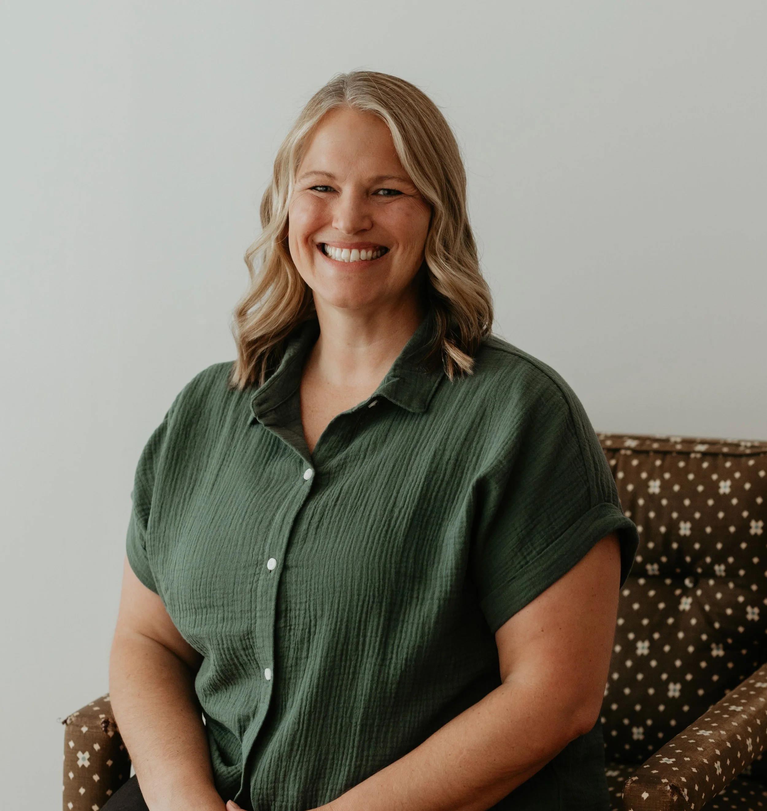 A Myofascial Release therapist with shoulder-length blonde hair, smiling, wearing a dark green button-up shirt, sitting on a patterned brown chair with white flowers, against a plain light wall background.