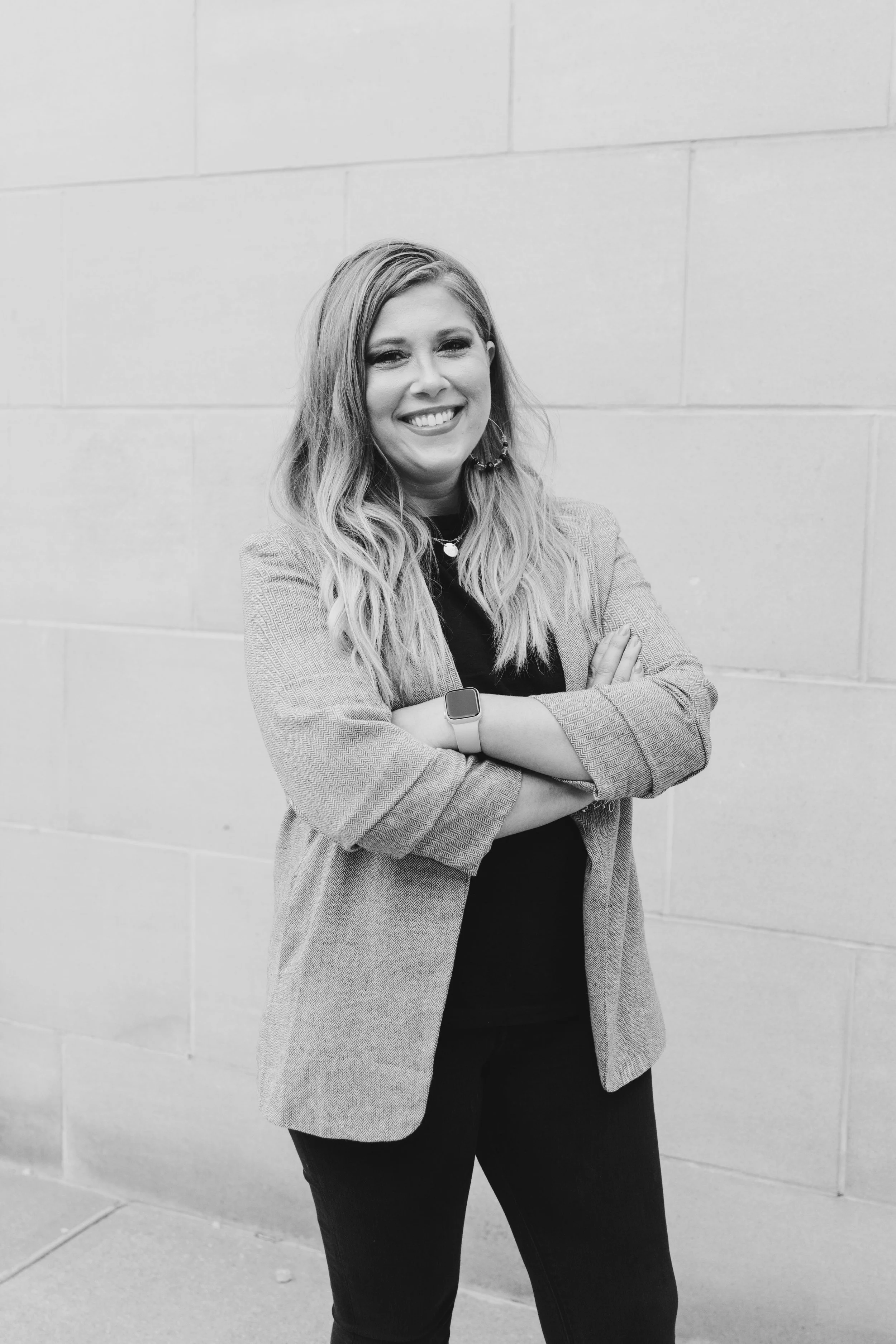 Black and white photo of a smiling woman with long wavy hair, standing against a brick wall, arms crossed, wearing a blazer, earrings, and a smartwatch.