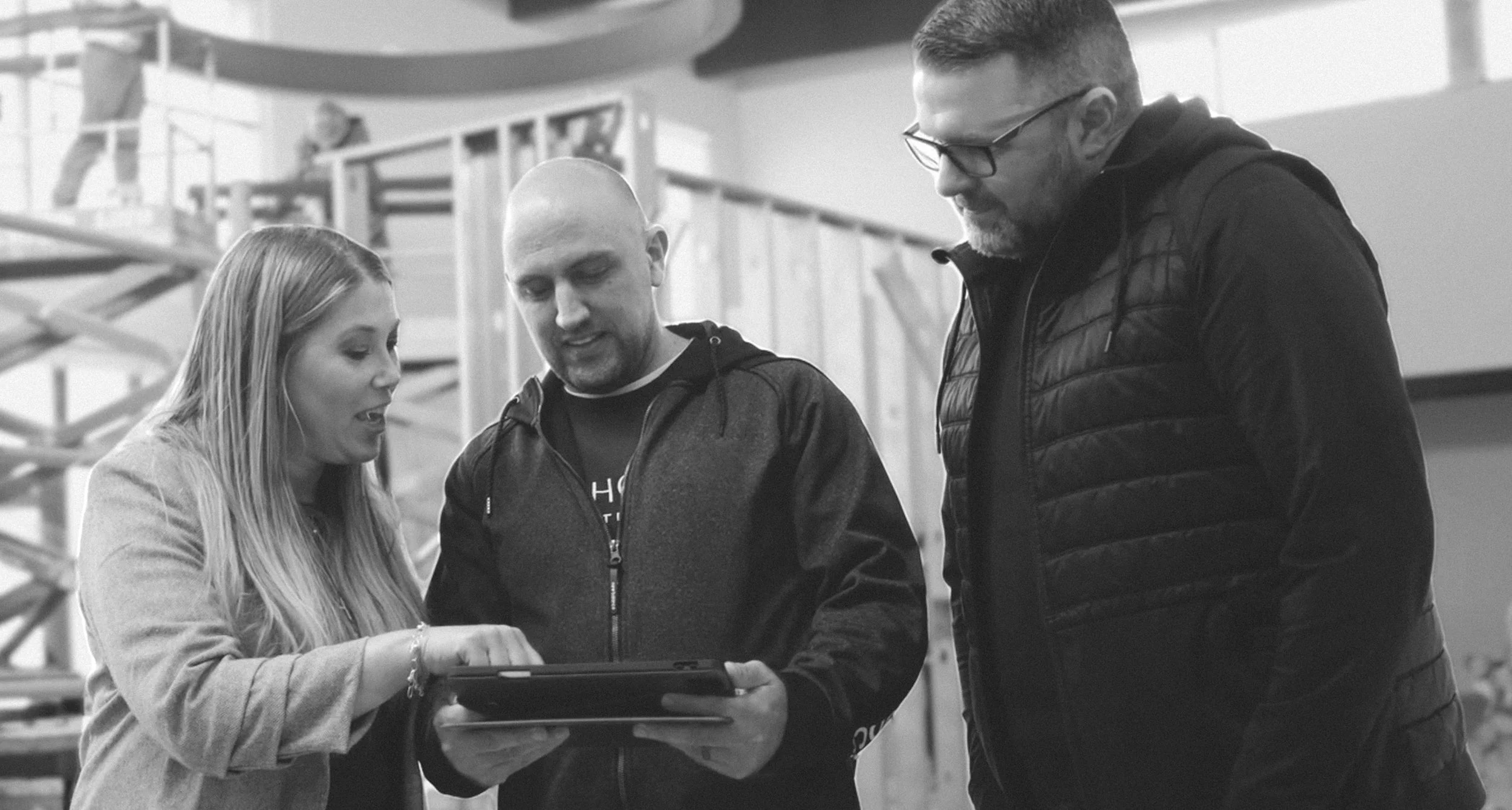 Three people, two men and a woman, are gathered around a tablet or small electronic device, looking at it together indoors in what appears to be a gym or recreational facility, with a climbing structure in the background.