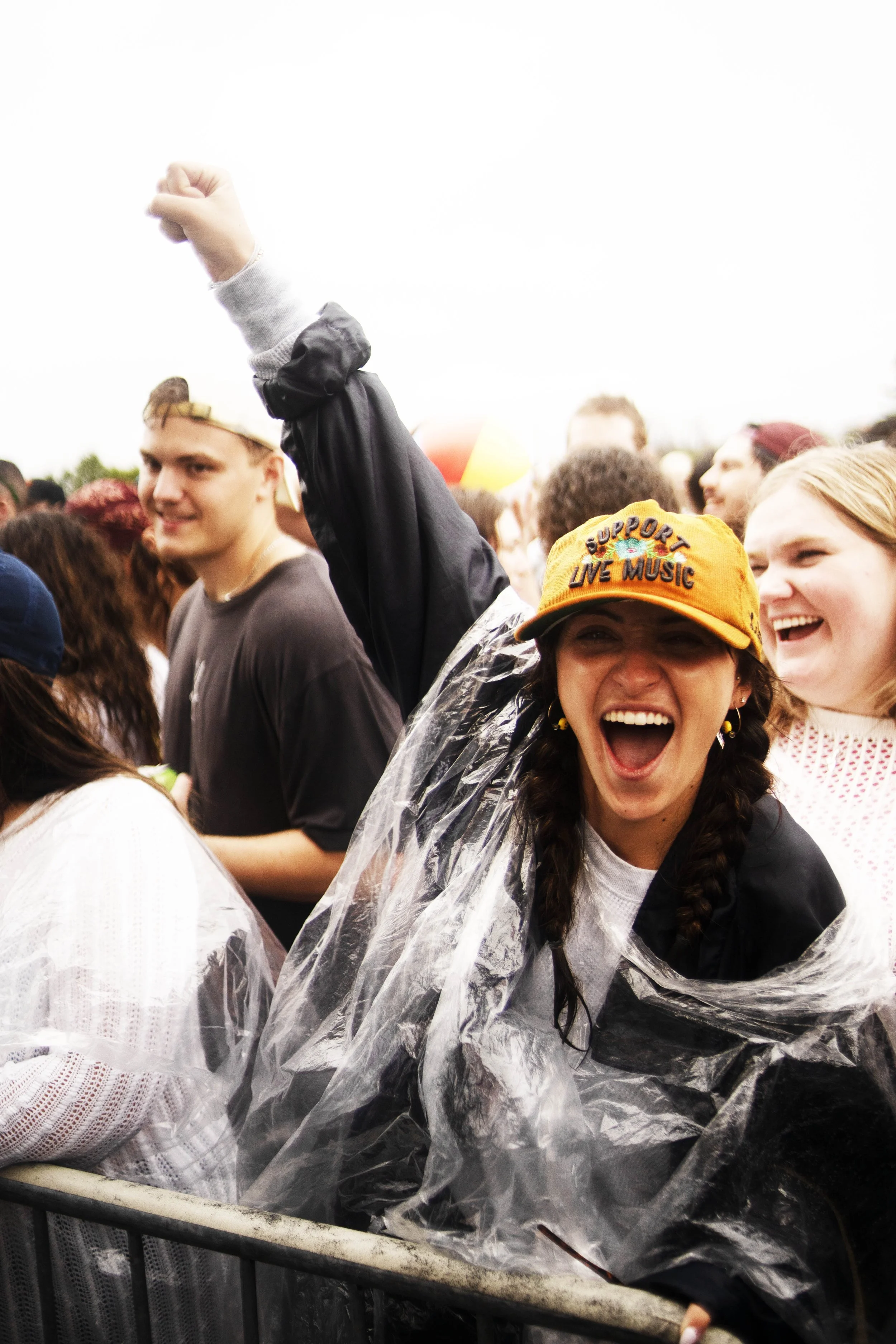Crowd of people at a music event, some people smiling and cheering, one woman in the foreground wearing a yellow cap that says 'Support Live Music' and a clear rain poncho.