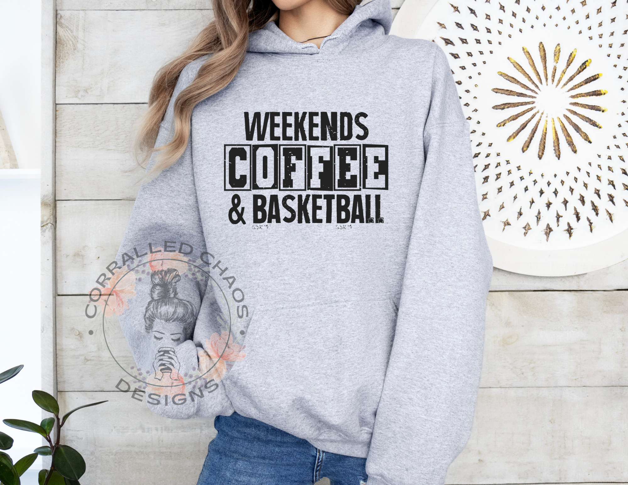 A woman wearing a gray hoodie with the text "Weekends Coffee & Basketball" in black lettering. The background includes textured white wood panels, a decorative art piece with a white and gold pattern, and a green plant with round leaves.