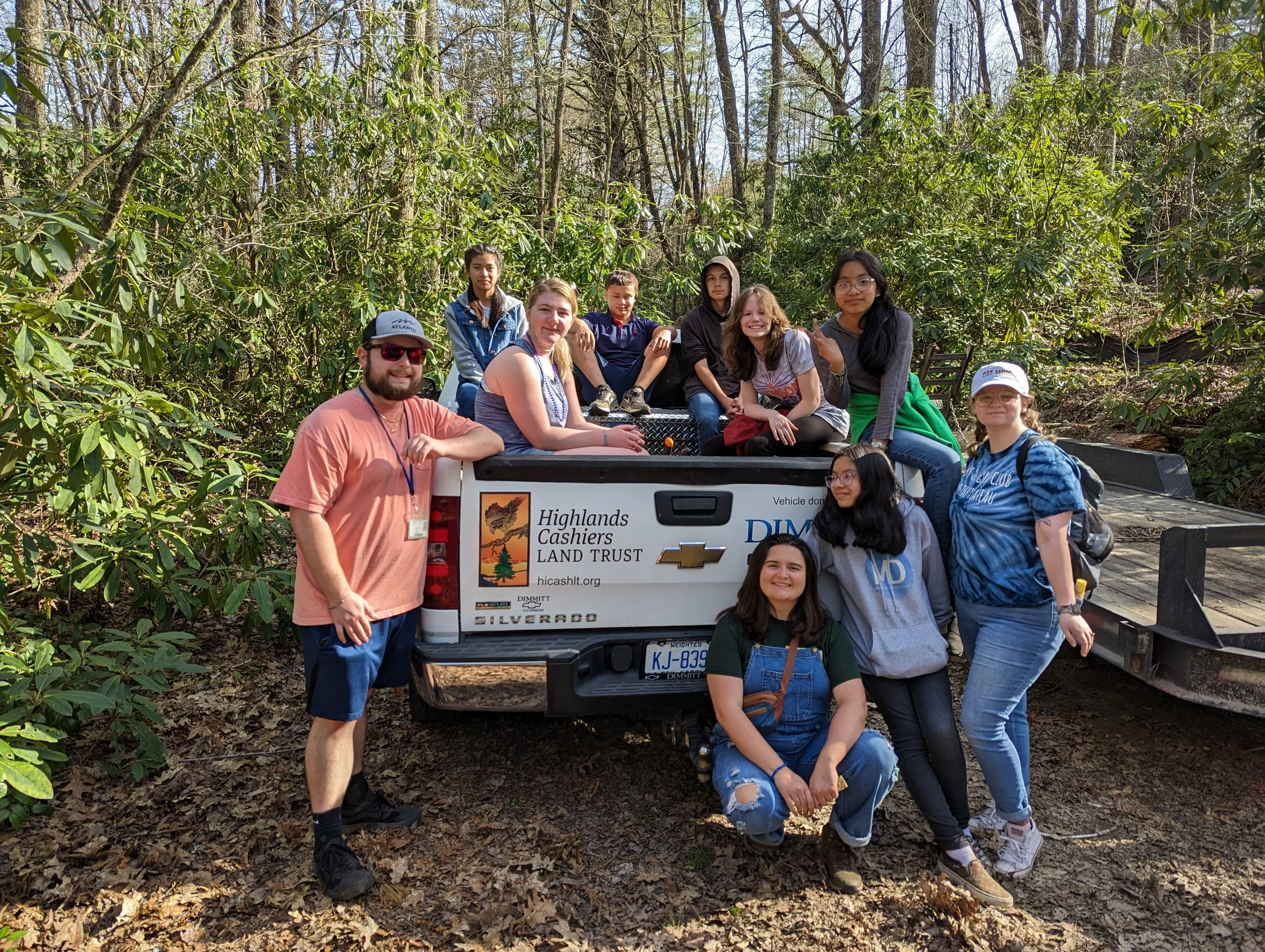 Group of eleven people, including children and adults, sitting and standing on the back of a Chevrolet Silverado pickup truck in a wooded area during daytime. The truck has organization stickers including Highlands Cashiers Land Trust and Dimmitt Chevrolet.