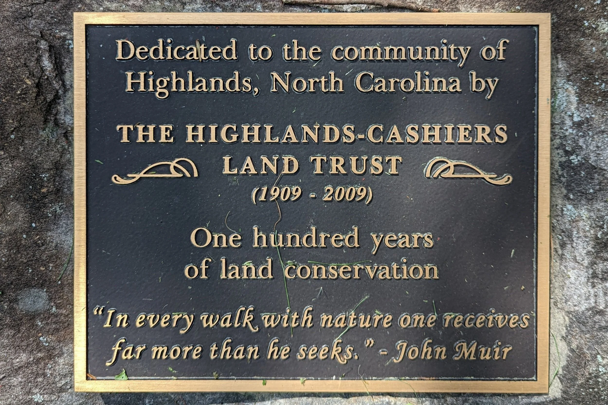 A bronze plaque dedicated to the Highlands Cashiers Land Trust, mounted on a rock, with engraved text honoring 100 years of land conservation from 1909 to 2009, including a quote by John Muir.