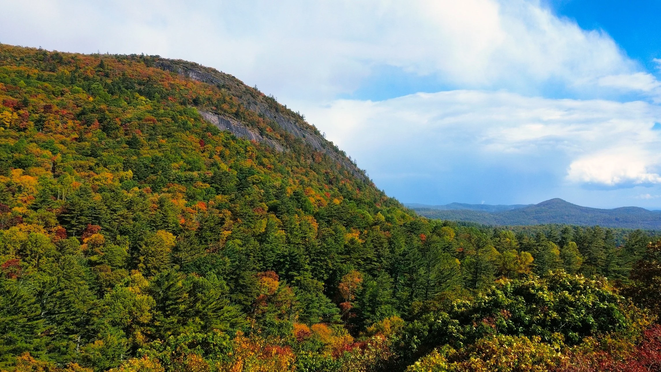 A scenic view of a mountain covered in dense green forest with patches of red, orange, and yellow fall foliage, under a partly cloudy sky.