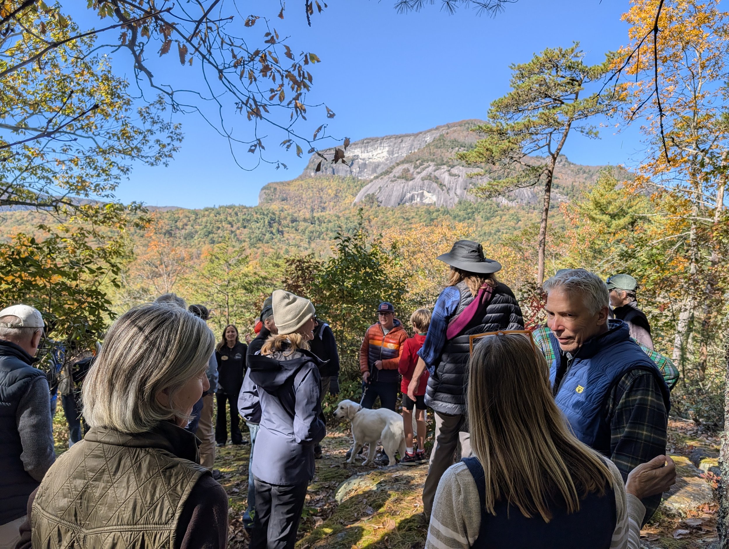 A group of people gathered outdoors on a forested trail during autumn with colorful fall leaves, mountain scenery, and a bright blue sky in the background.