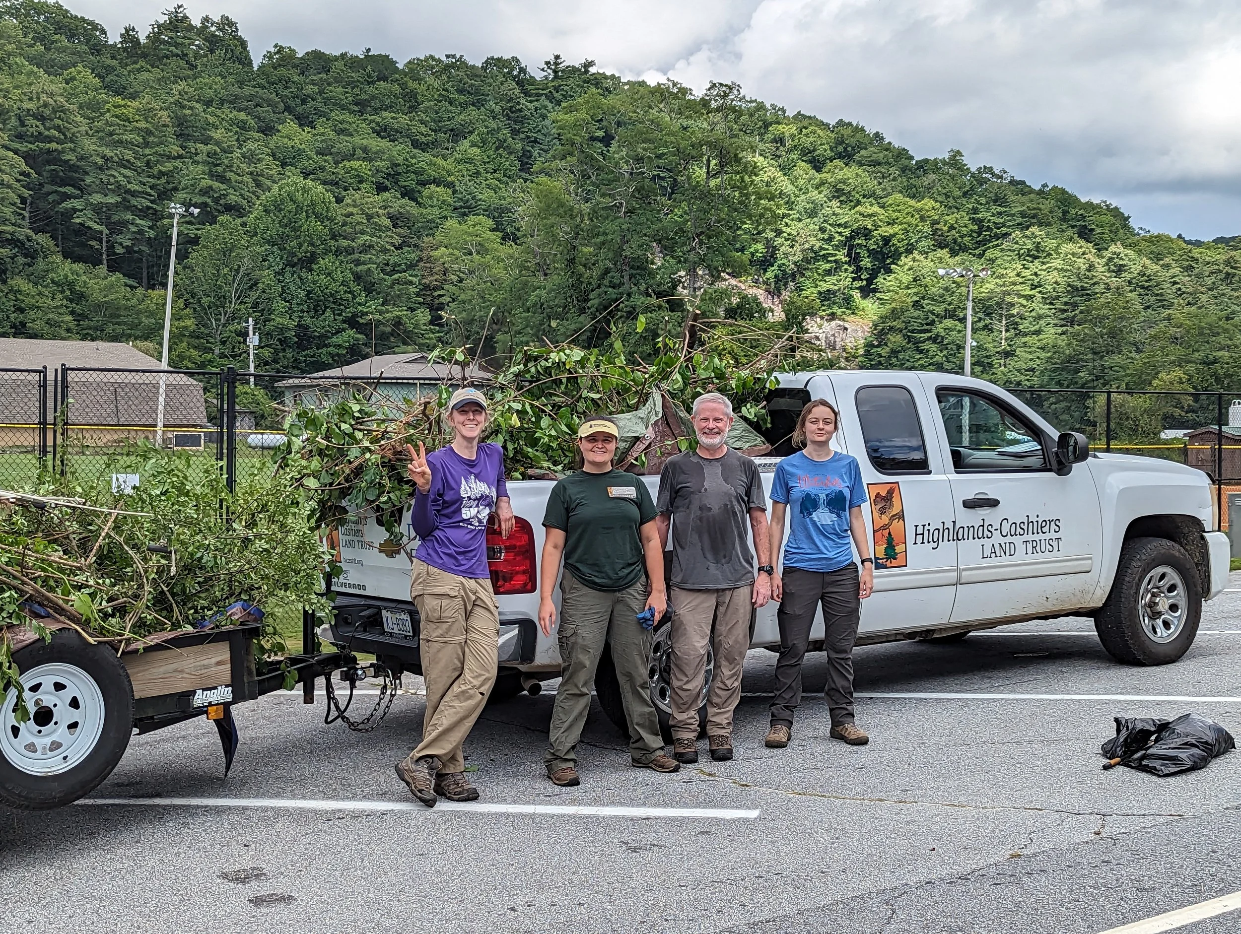 Four smiling people standing next to a white Highlands Cashiers Land Trust pickup truck and a small trailer filled with greenery on a parking lot, with a green forested hill and cloudy sky in the background.