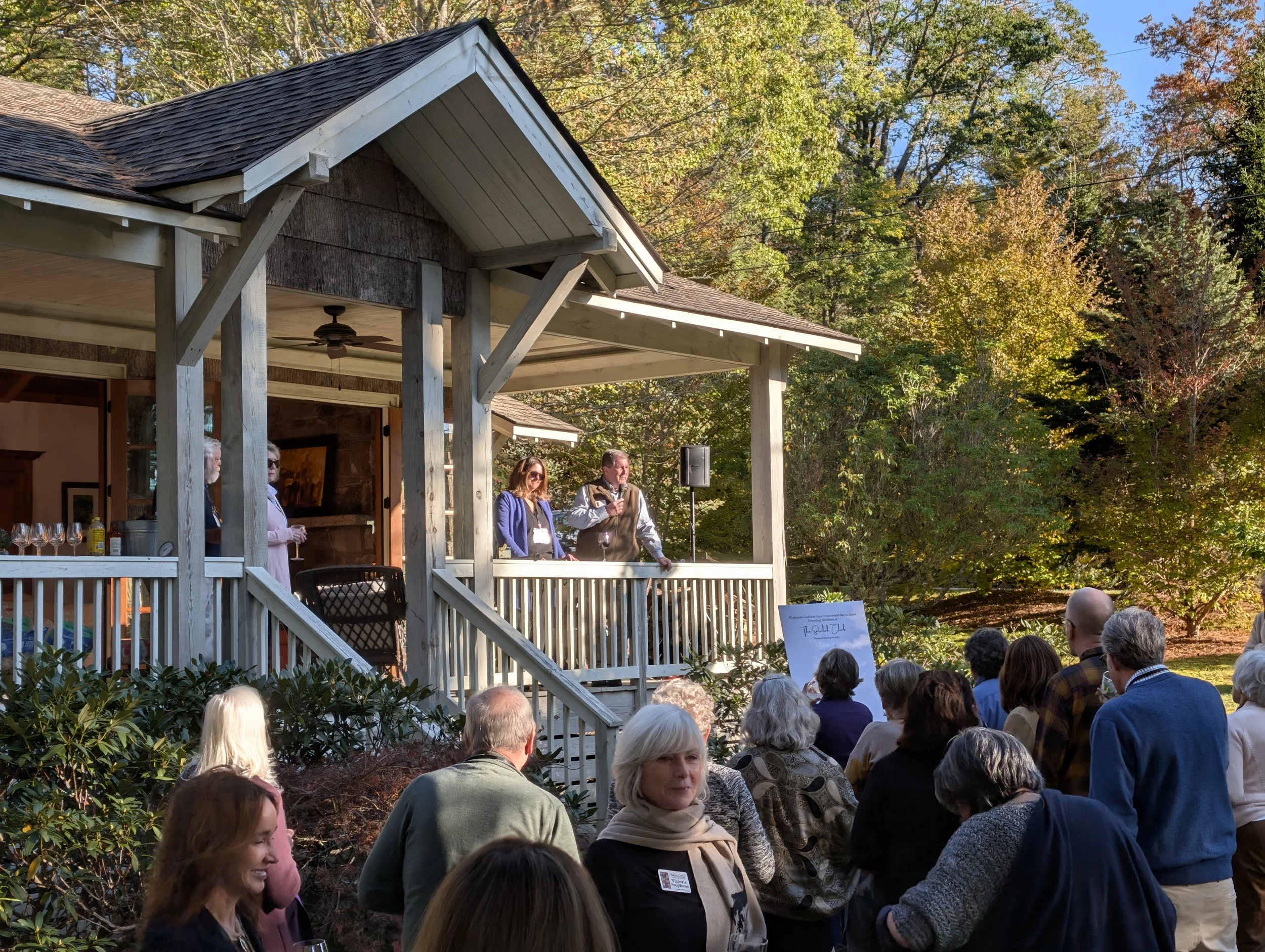 A gathering of people on a porch and lawn during an outdoor event in the daytime, with a speaker addressing the crowd from the porch surrounded by trees with fall foliage.
