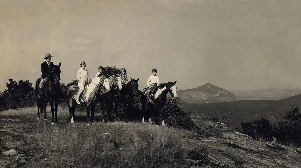 Six people riding horses in a natural landscape with rolling hills in the background, black-and-white photo.