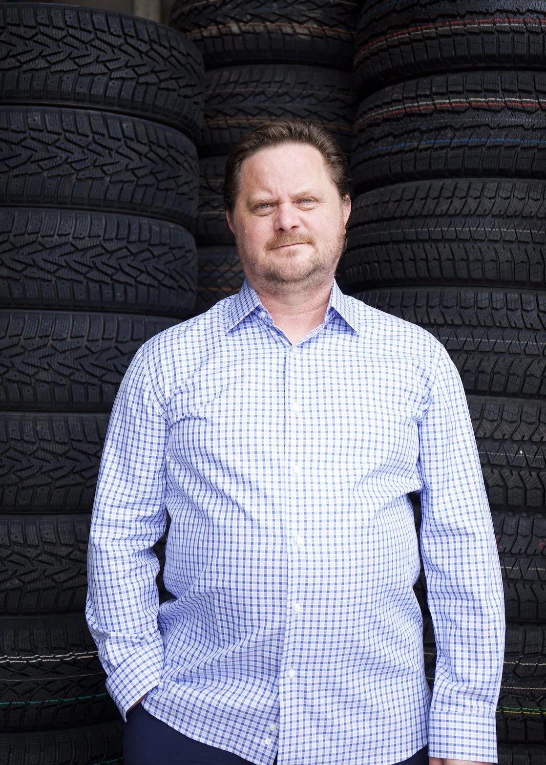 A man with a beard in a checkered shirt standing in front of stacking tires.