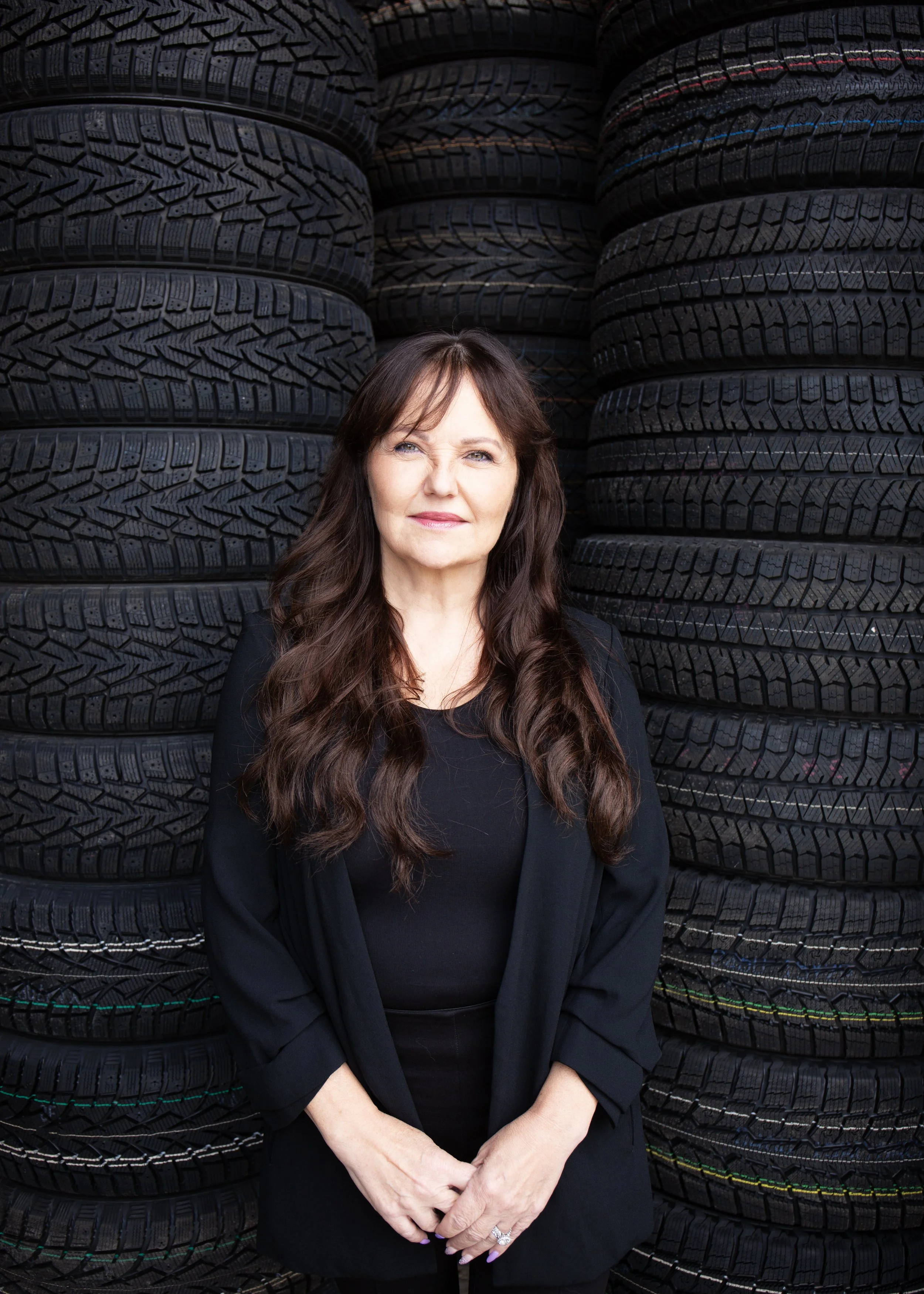 A woman with long brown hair standing in front of stacked black tires, winking, and smiling slightly.