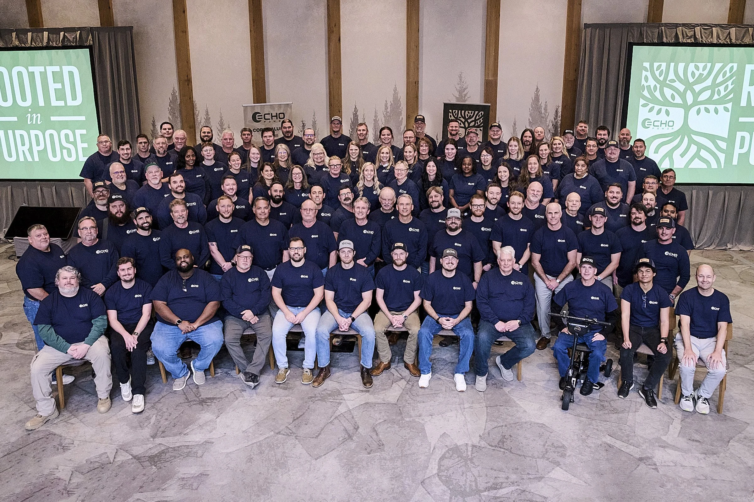 A large group of people, mostly wearing dark blue T-shirts with a logo, gathered in a conference room for a group photo. Two large screens with green and white logos are visible on either side, with one reading 'Rooted in Purpose'.