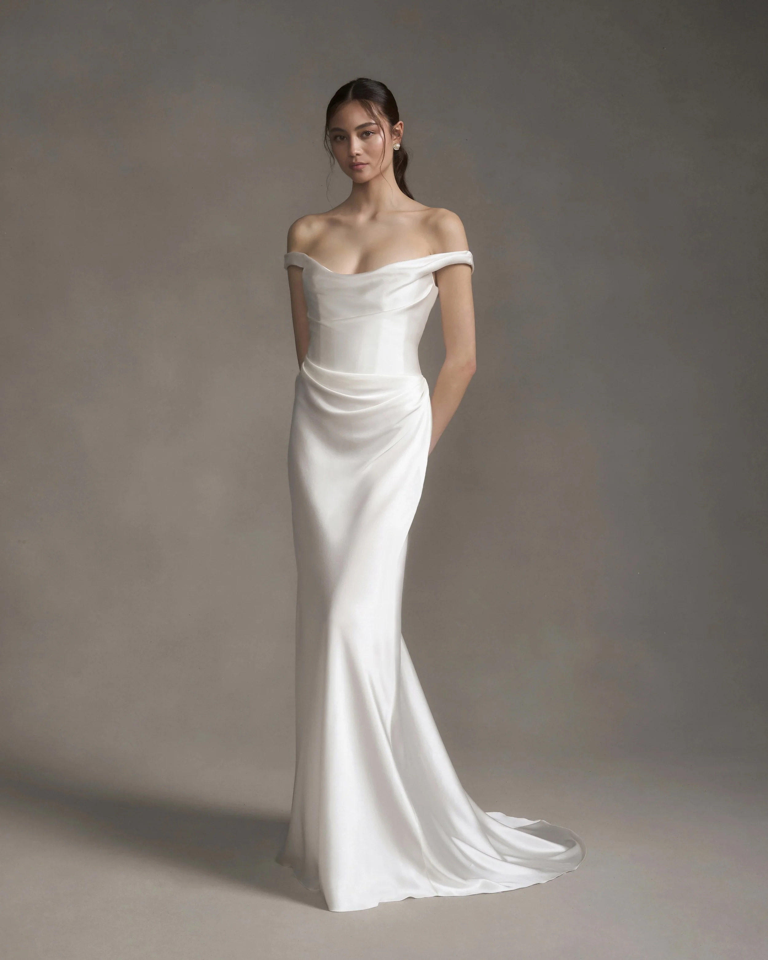 Model wearing an off-the-shoulder ivory satin wedding gown with a softly draped bodice and skirt.