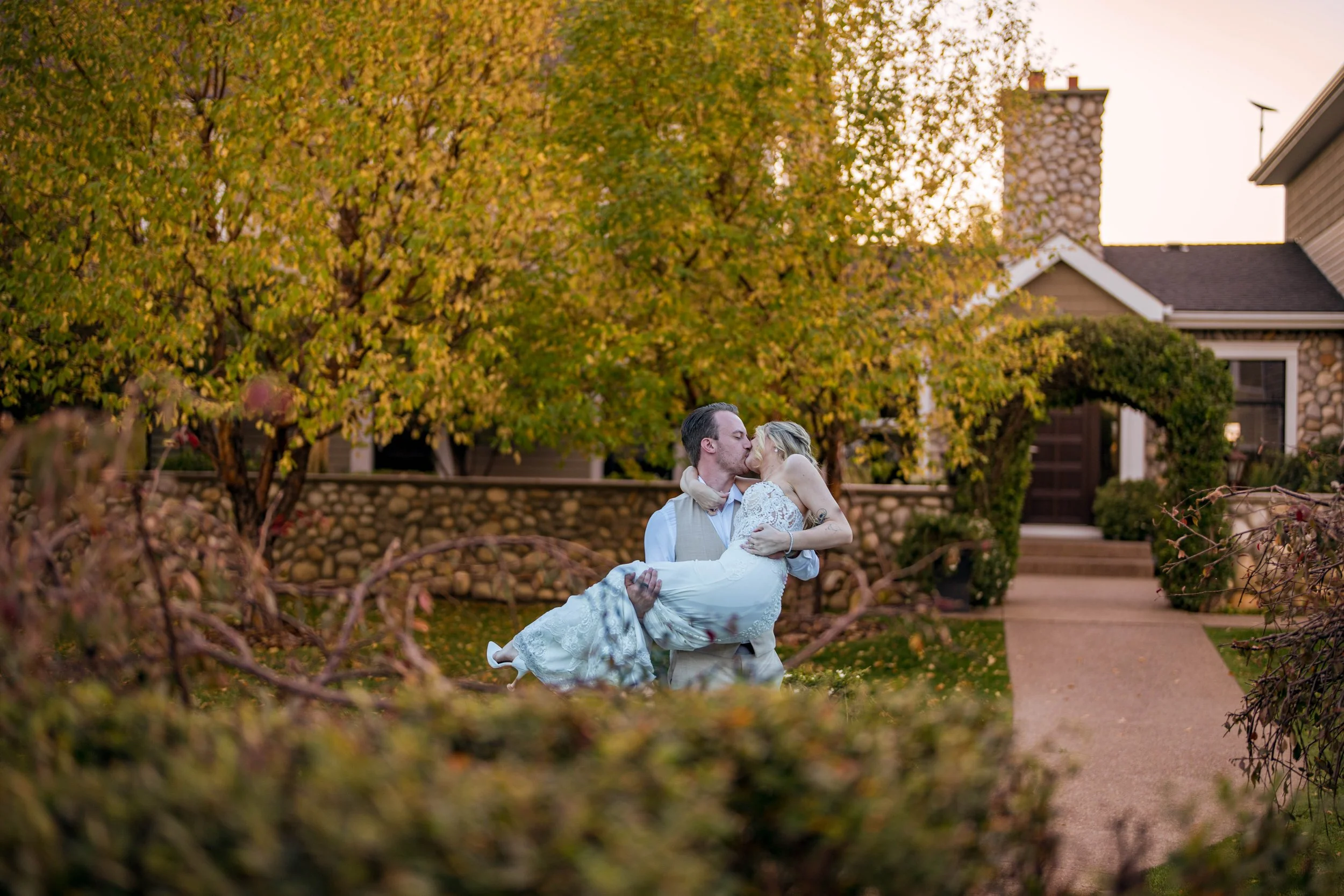 A couple dressed in wedding attire sharing a kiss outdoors during sunset, with trees and a house in the background.