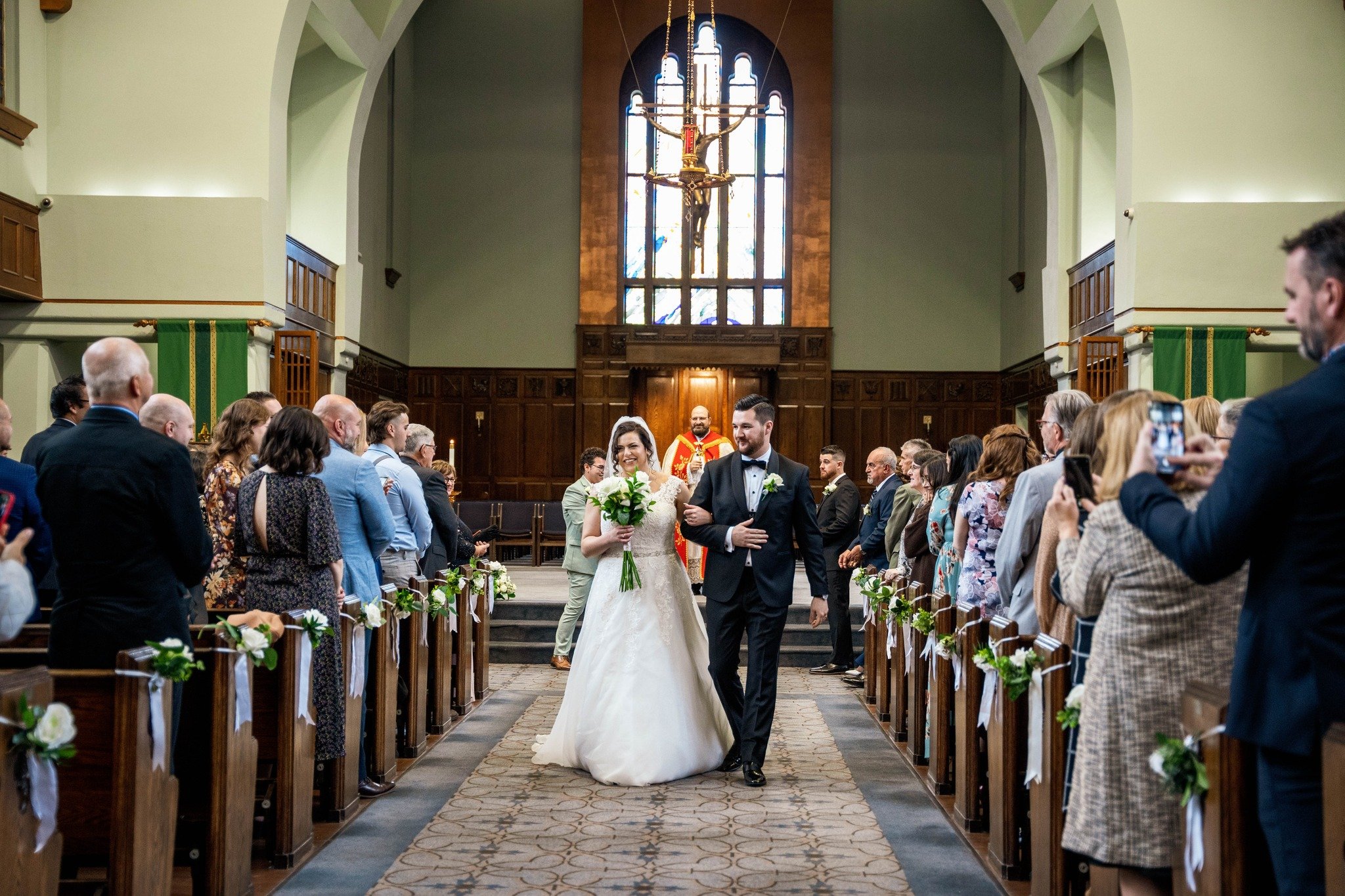 A bride and groom walking down the aisle of a church after their wedding ceremony. The crowd is standing and taking pictures, with a priest standing behind them. The church has wooden pews decorated with white flowers and ribbons, and a large stained
