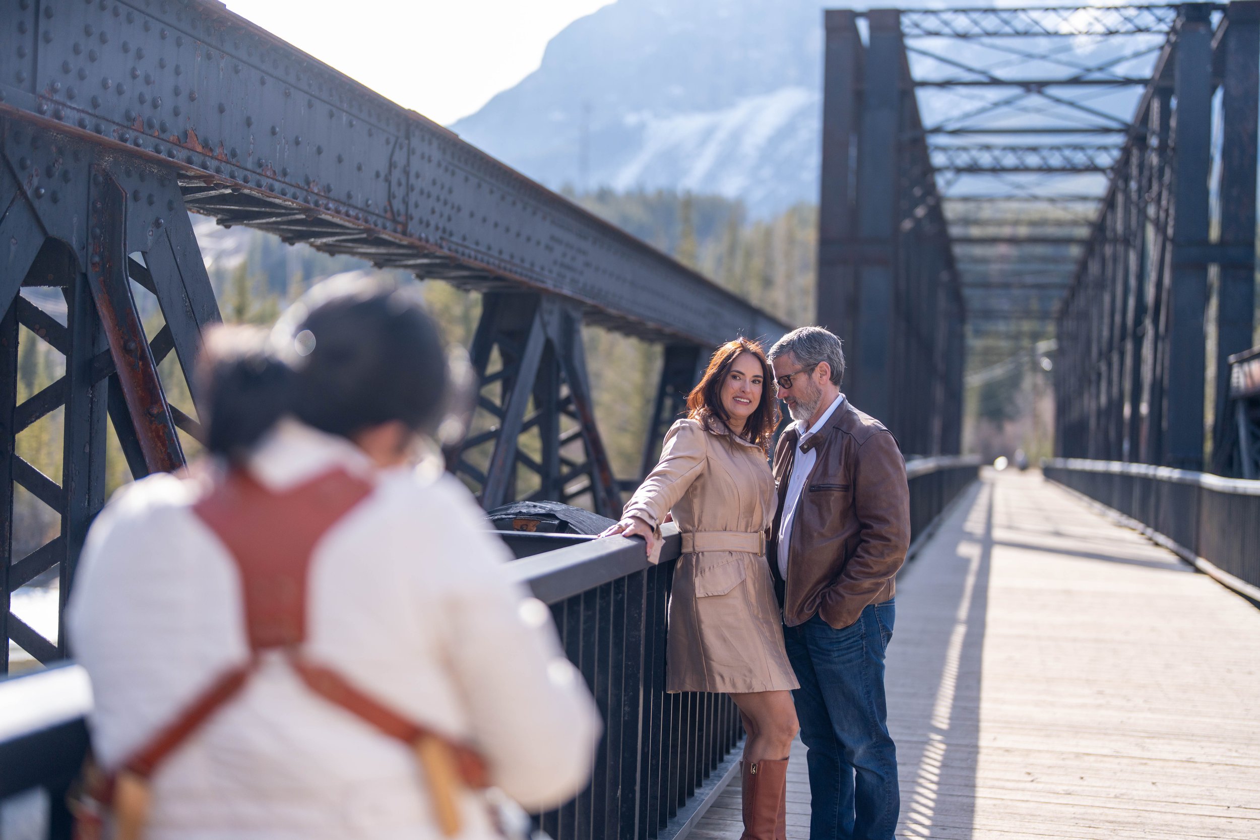 Two people, a woman in a beige coat and a man in a brown leather jacket, standing beside a bridge railing outdoors, with a mountain in the background. A third person with short dark hair and glasses, wearing a white shirt, is in the foreground with their back to the camera.