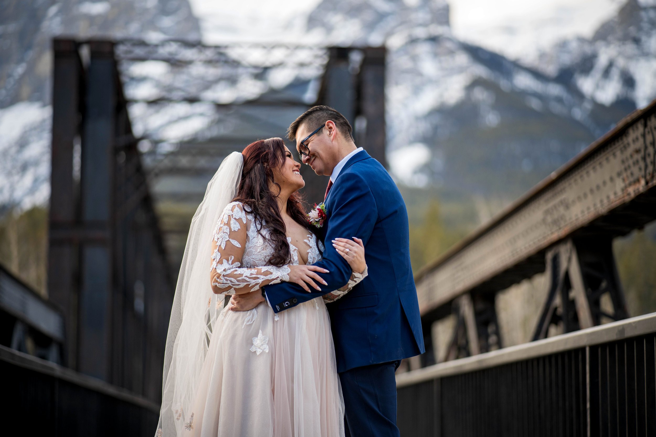 Bride and groom embracing outdoors with a mountain and a train bridge in the background.