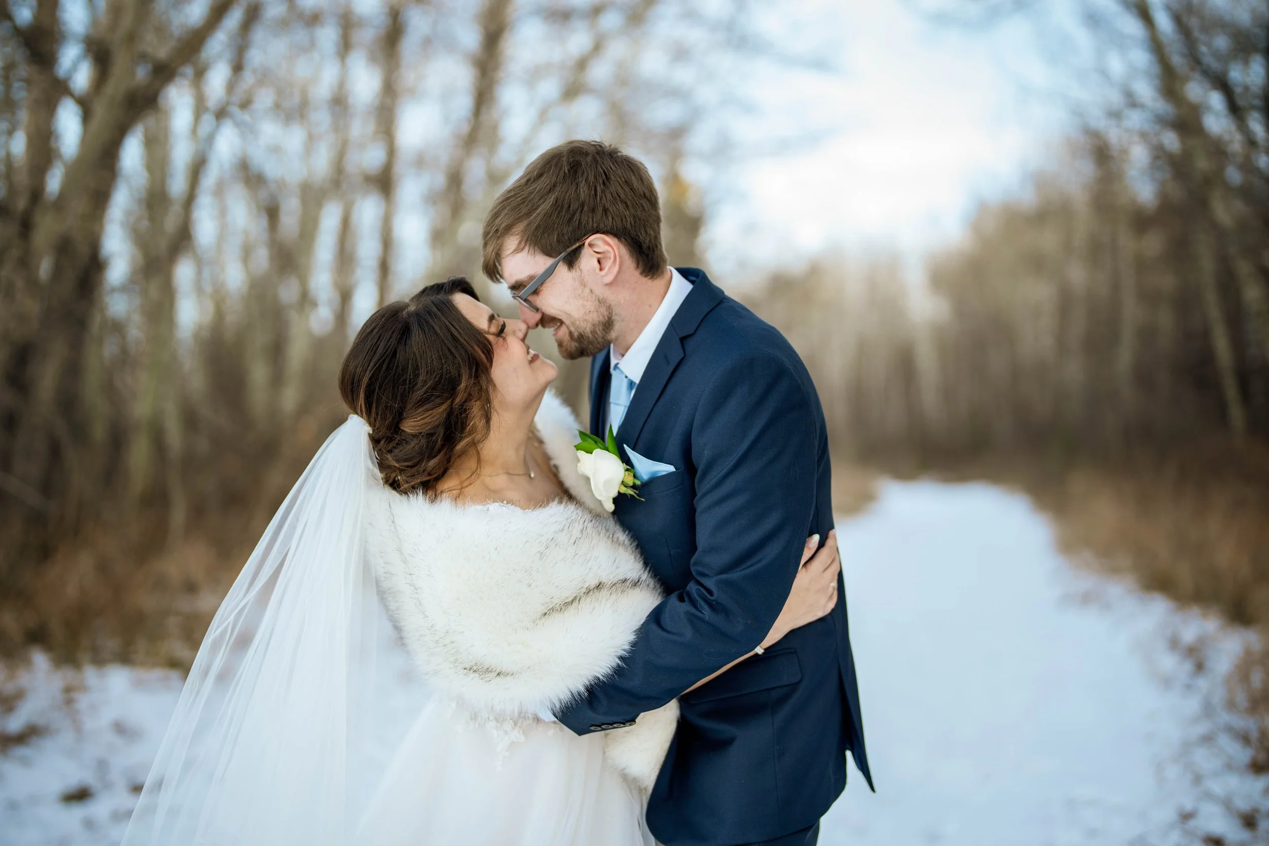 A newlywed couple embraces in a snowy outdoor setting with the woman in a wedding dress and fur shawl, and the man in a navy suit and glasses, nose to nose, surrounded by leafless trees.