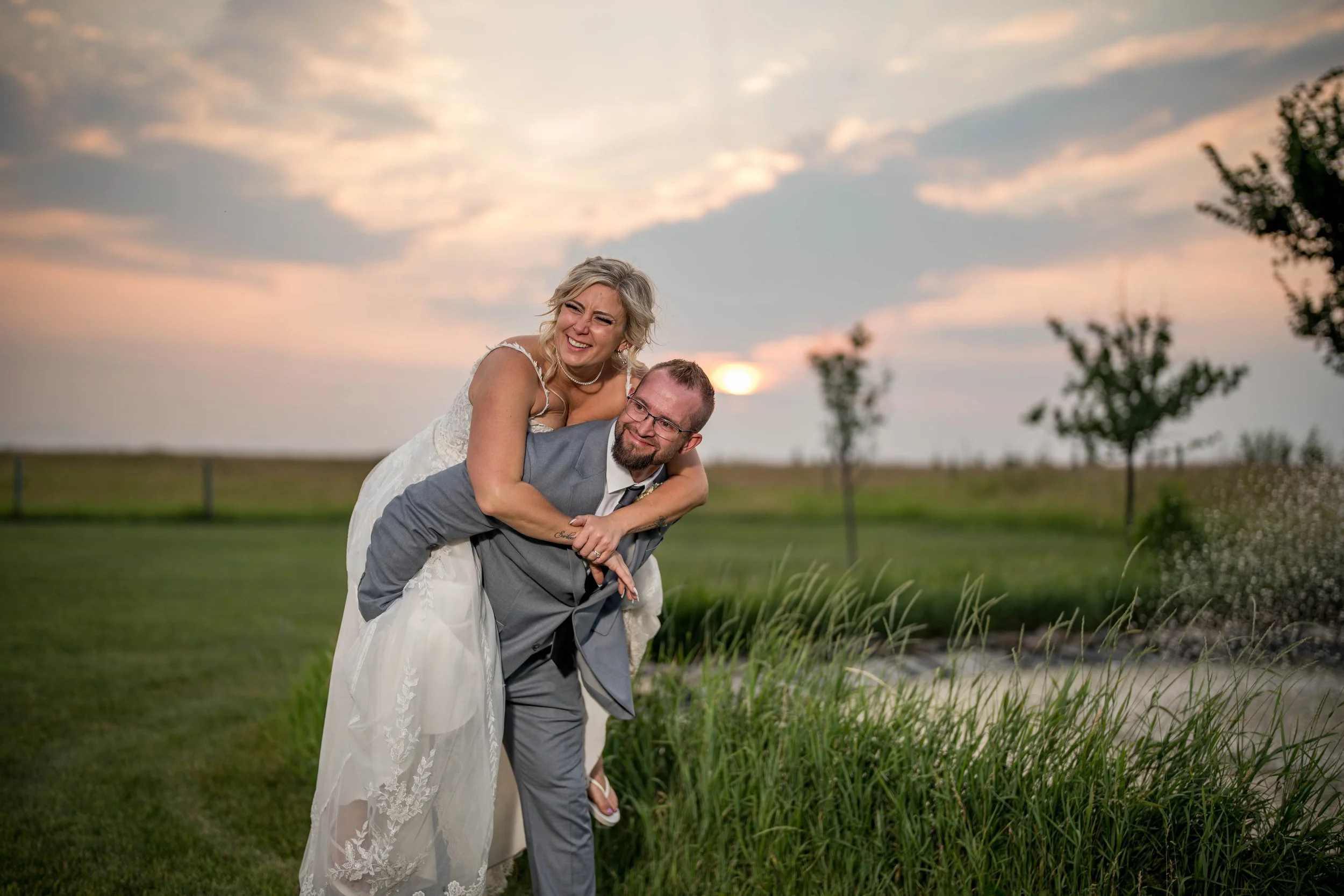 A bride and groom outdoors during sunset, with the bride on the groom's back, smiling and laughing on a grassy field with trees and a cloudy sky in the background.