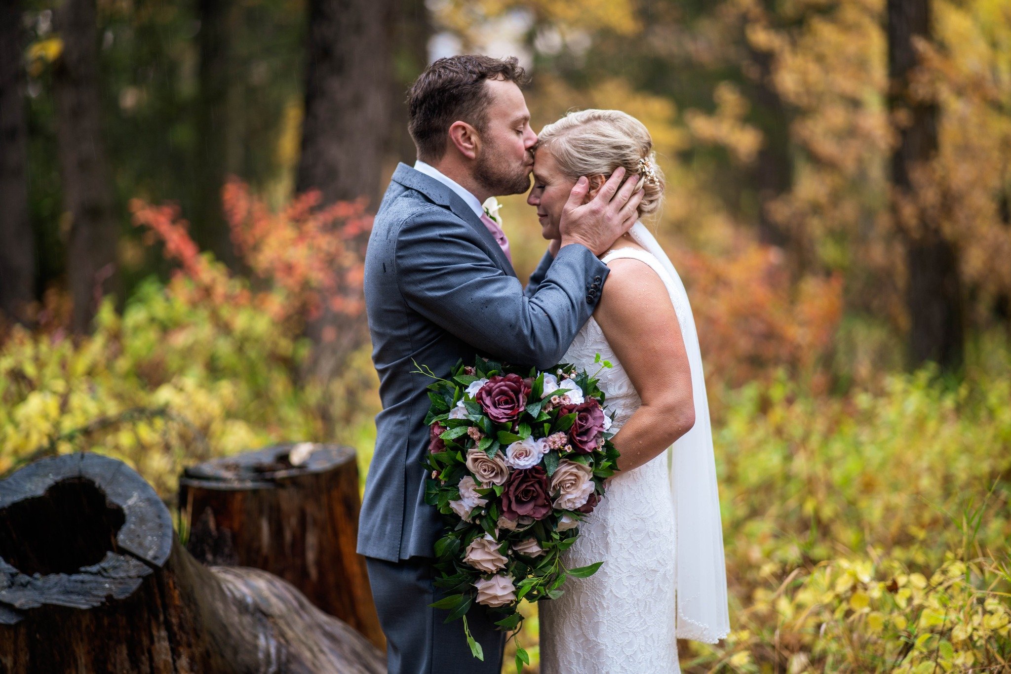 A groom kissing a bride on the forehead during a wedding, holding her face in his hands, in an autumn forest setting. The bride is holding a large bouquet of flowers with roses and greenery.