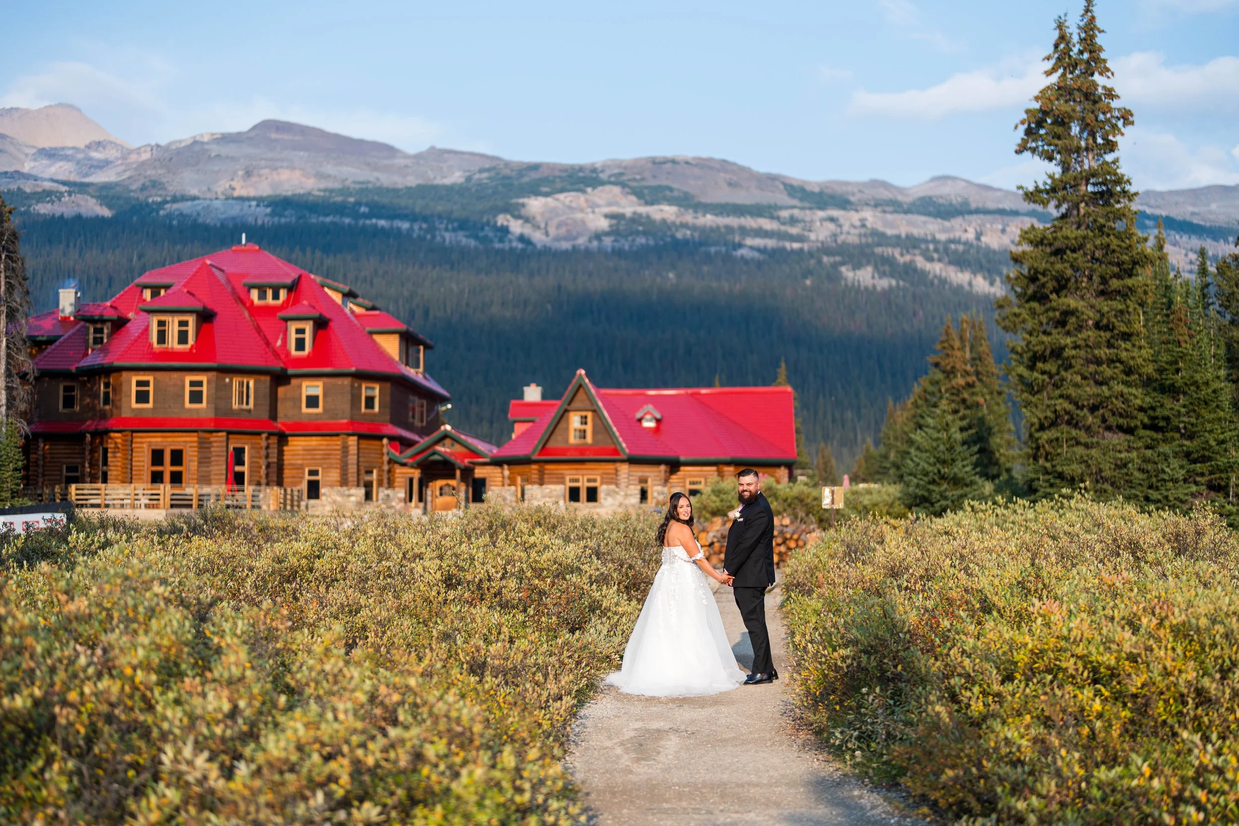 A bride and groom holding hands on a dirt path with a large mountain lodge in the background, surrounded by green trees and bushes, under a partly cloudy sky.