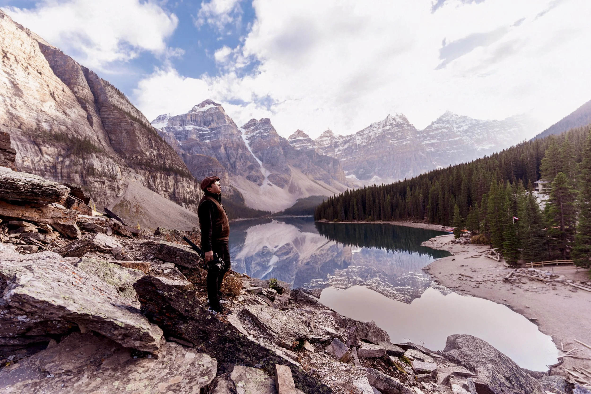 A person standing on rocky terrain beside a lake with snow-capped mountains and pine trees in the background.