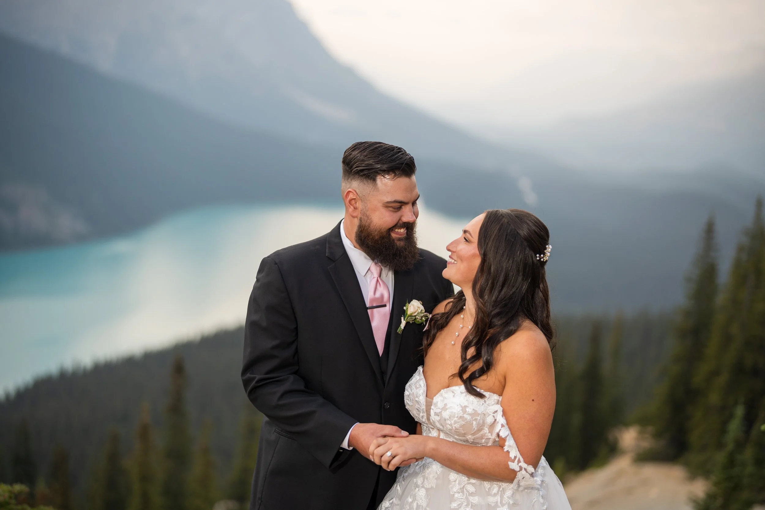 A newlywed couple in wedding attire standing outdoors near a mountain lake, holding hands and gazing into each other's eyes with a forest and mountains in the background.