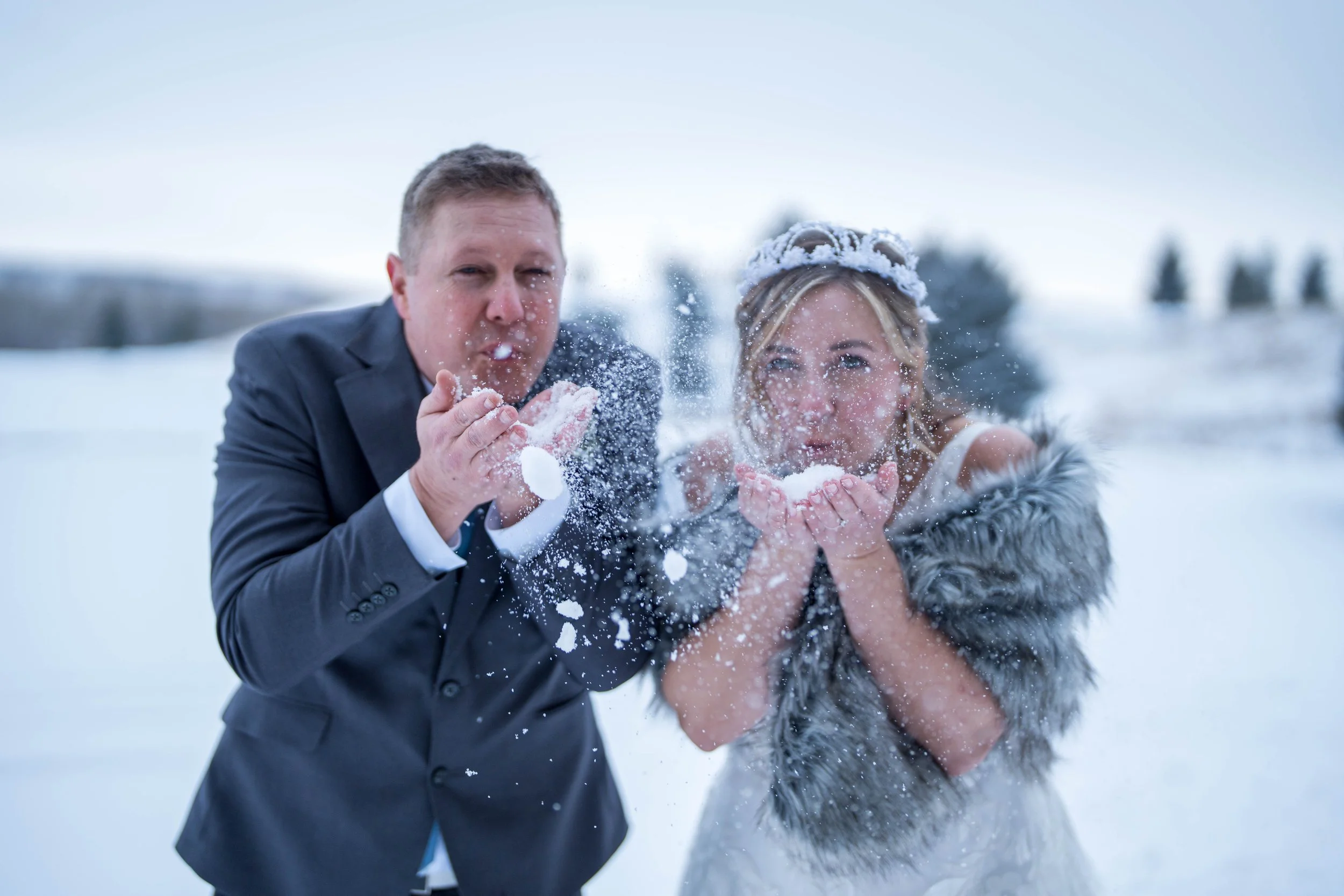 A man in a suit and a woman in a dress blowing snow towards the camera in a snowy outdoor winter landscape.