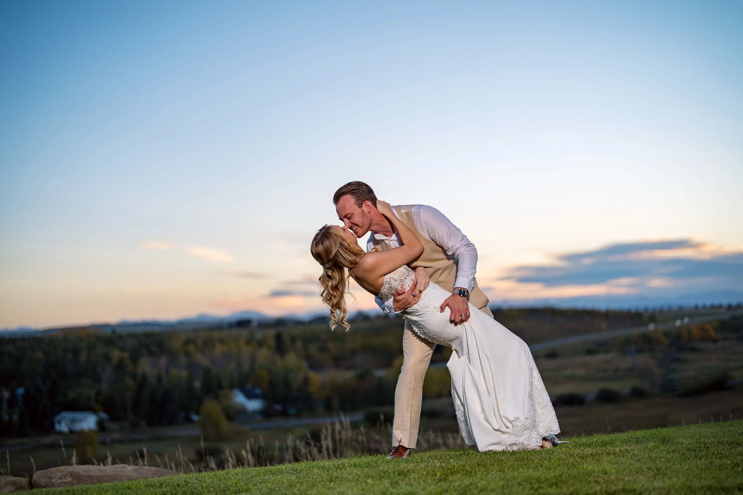 A newlywed couple dancing outdoors on a grassy hill at sunset, with a scenic landscape and sky in the background.
