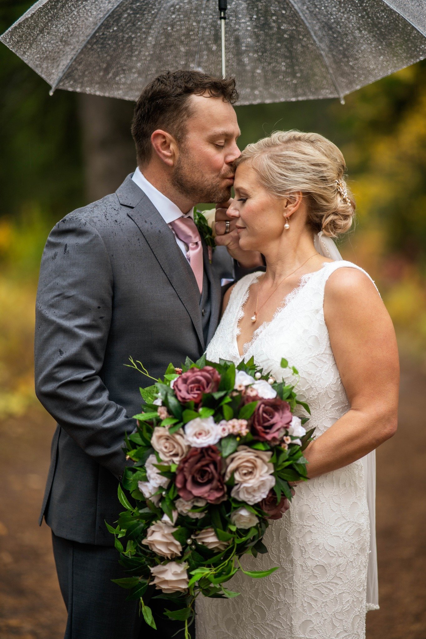A bride and groom standing close together outdoors under a transparent umbrella. The groom is kissing the bride's forehead while she holds a large bouquet of pink, white, and purple roses. The scene appears to be during a wedding, with the couple dressed in wedding attire and surrounded by blurred autumn trees.