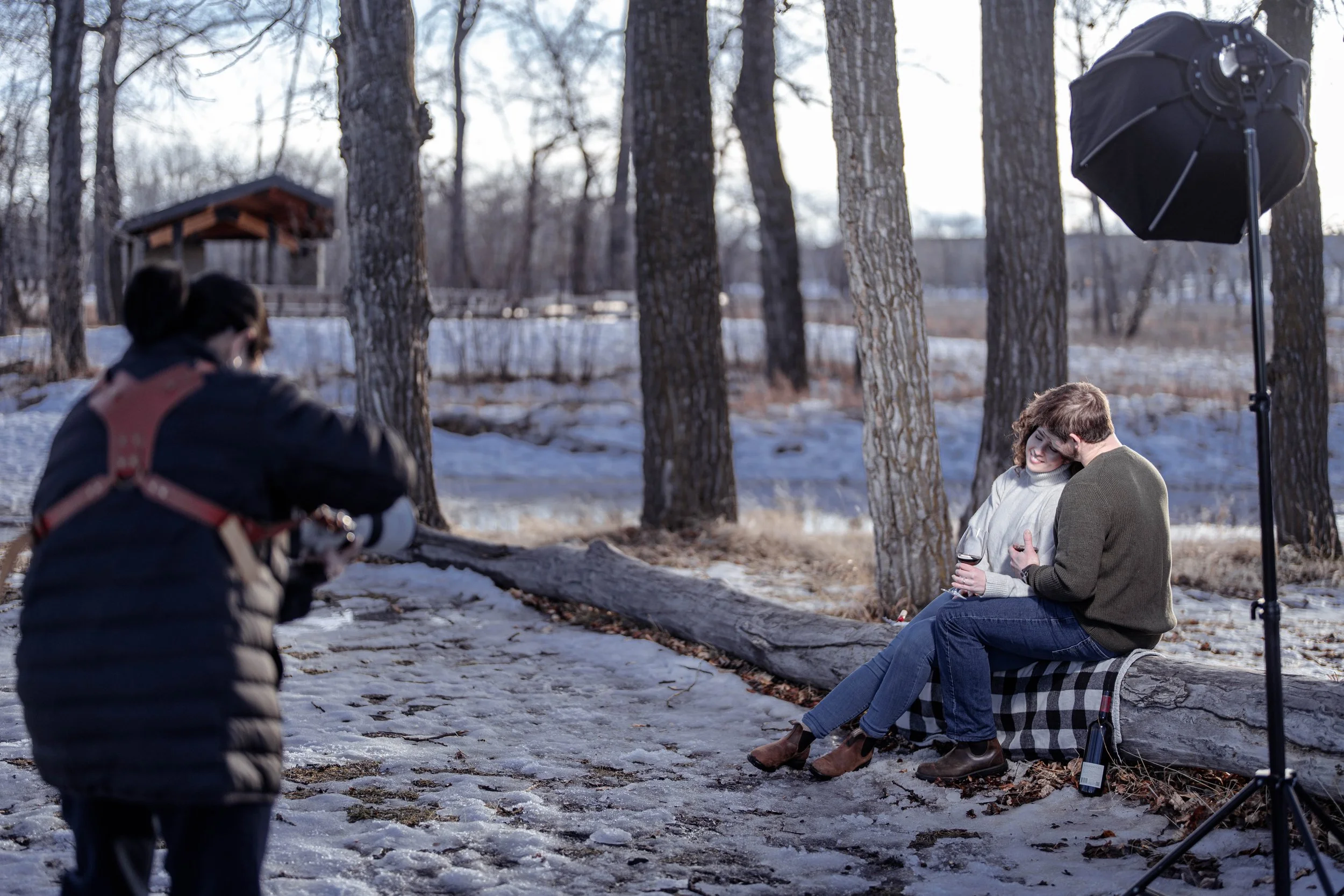 A couple sitting on a log in a snowy forest doing a photoshoot, with a photographer and lighting equipment nearby.