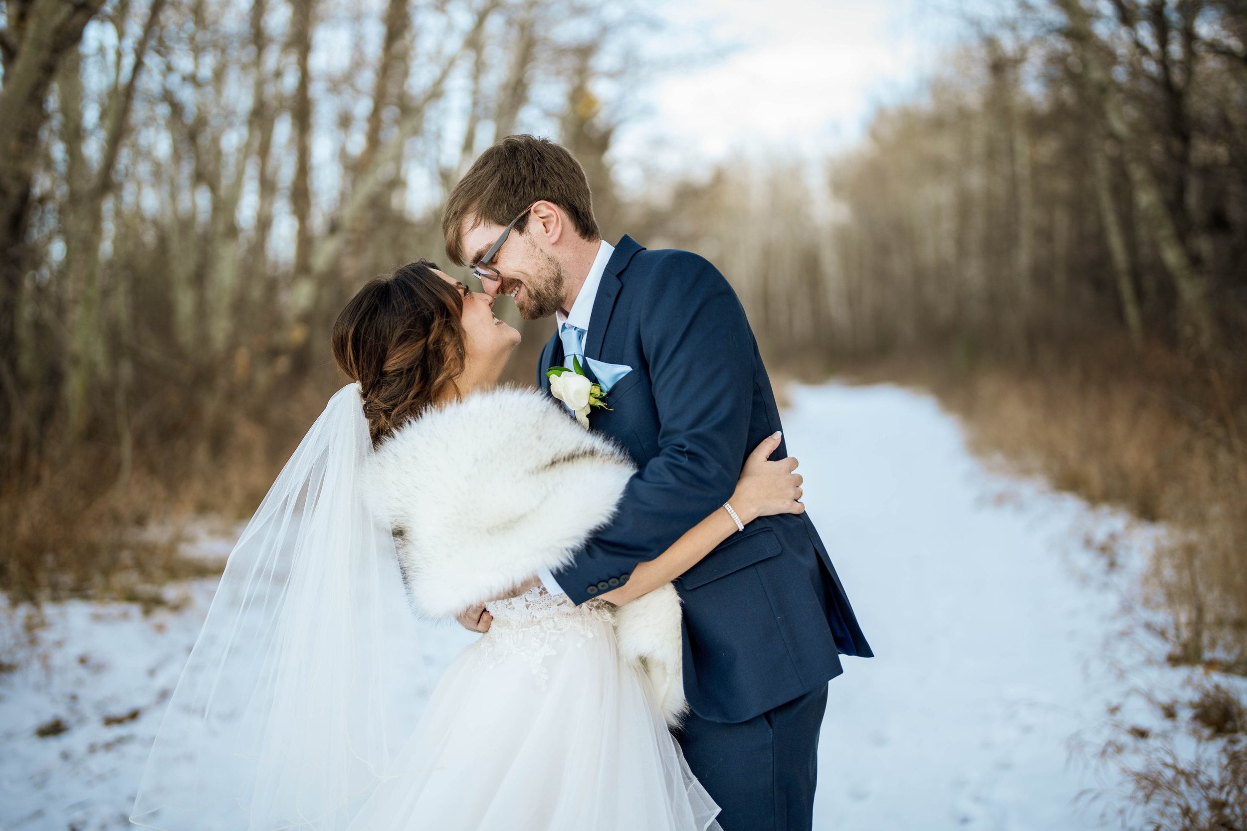 A bride and groom embracing outdoors on a snowy winter day, with trees in the background.