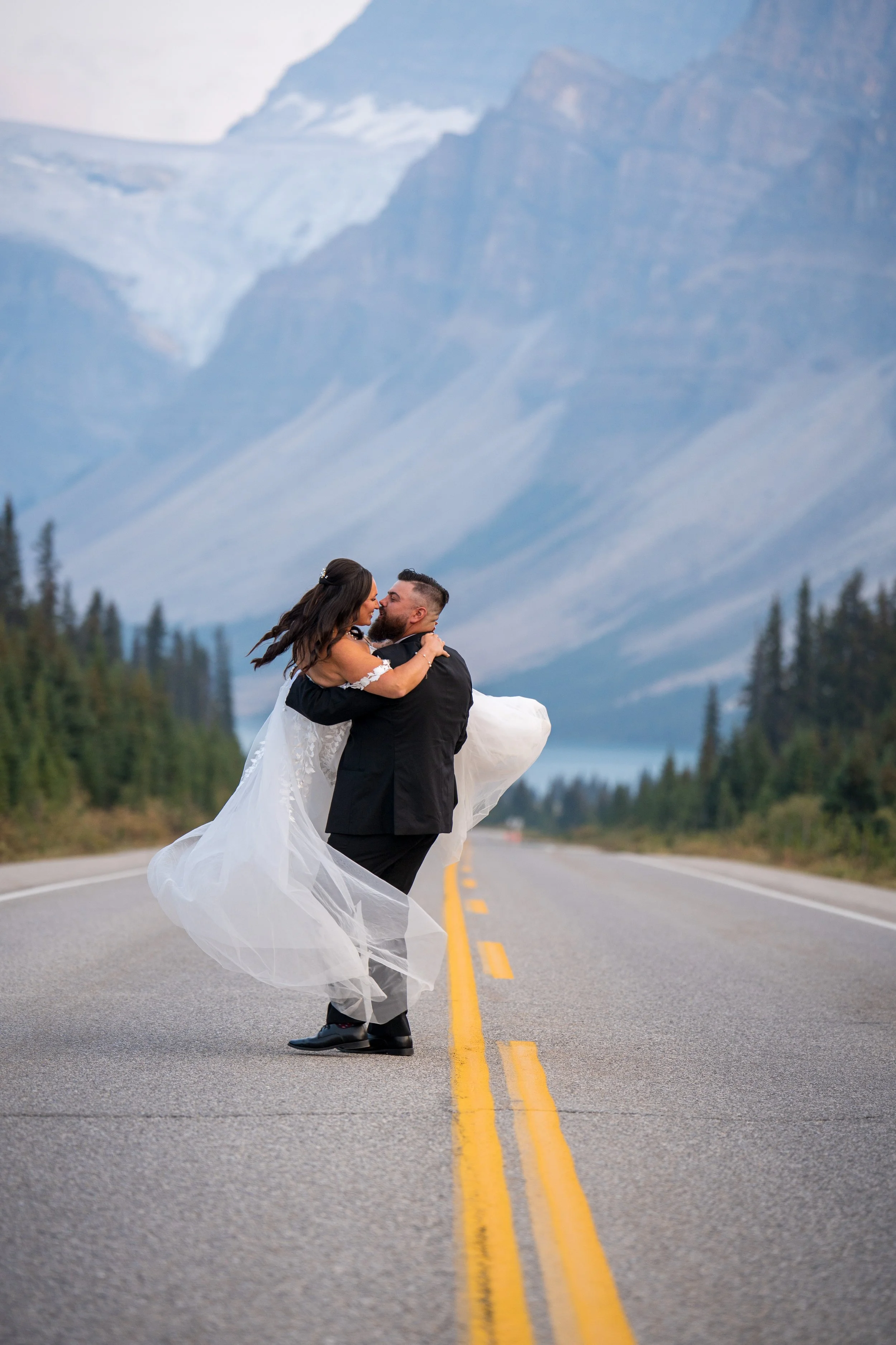 A newlywed couple kissing on an empty mountain road with scenic mountain scenery in the background.