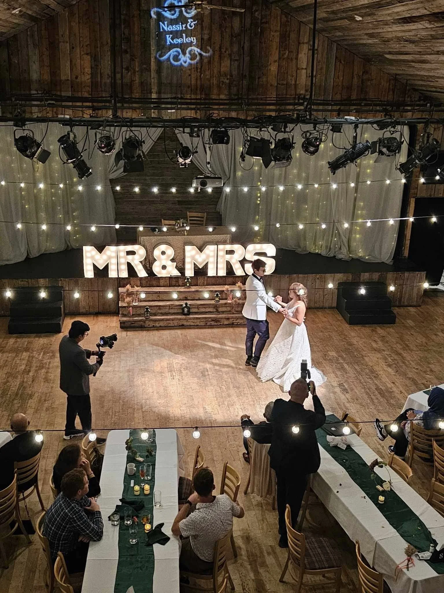 A bride and groom dancing on a wooden floor at their wedding, with guests seated at tables watching. Large illuminated letters spelling "MR & MRS" are on a stage behind them, decorated with string lights. The setting has rustic wood walls and ceiling, with lighting and decoration suitable for a wedding reception.