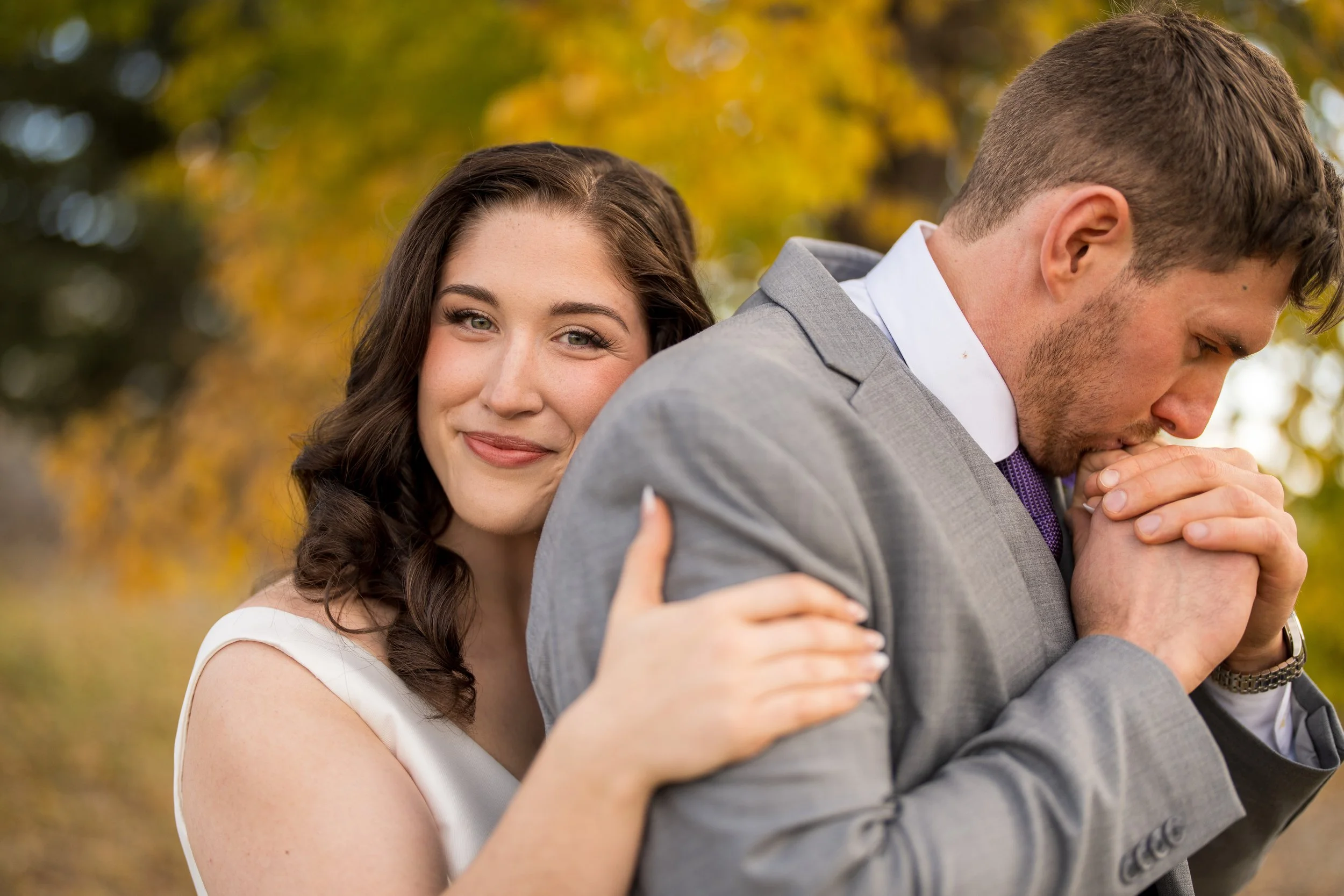 A woman with long brown hair smiling and touching a man in a gray suit, who is kissing his hands in prayer, outdoors with autumn leaves in the background.