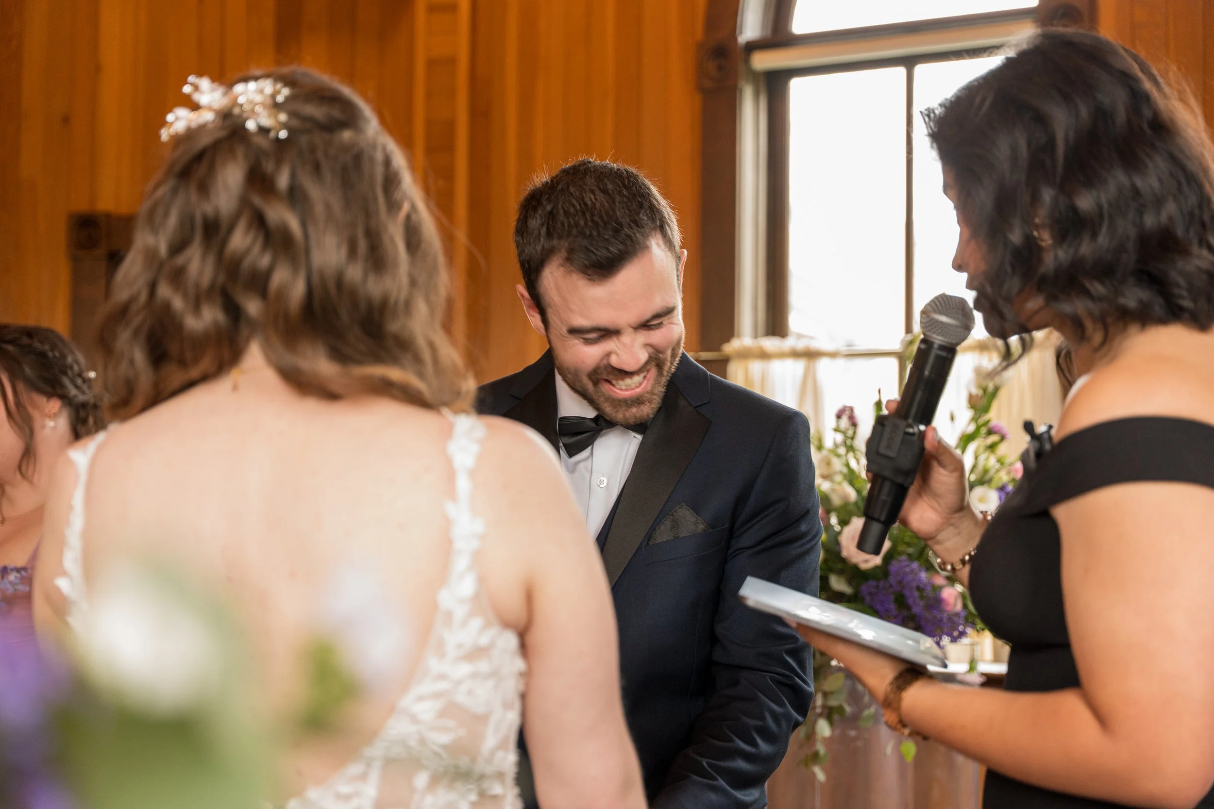 A wedding ceremony with a bride and groom smiling at each other while a woman holding a microphone and reading from a tablet officiates.